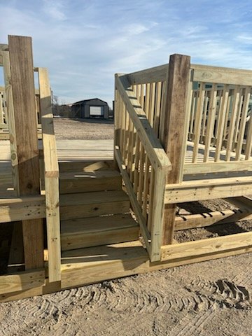 New wooden deck in construction on a dirt lot with a small shed and cloudy sky in the background.