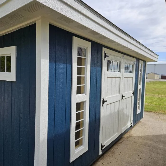 A small modern blue shed with white trim, windows, and double doors, situated on a grassy area under a cloudy sky.