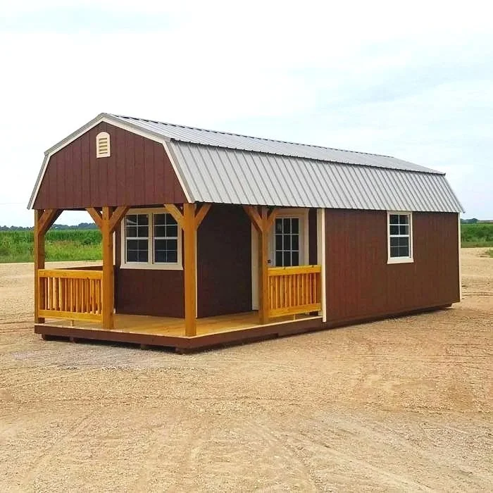 A small, brown storage shed with a metal roof, a porch with yellow wooden railings, and three windows, set on a dirt lot with green fields in the background.