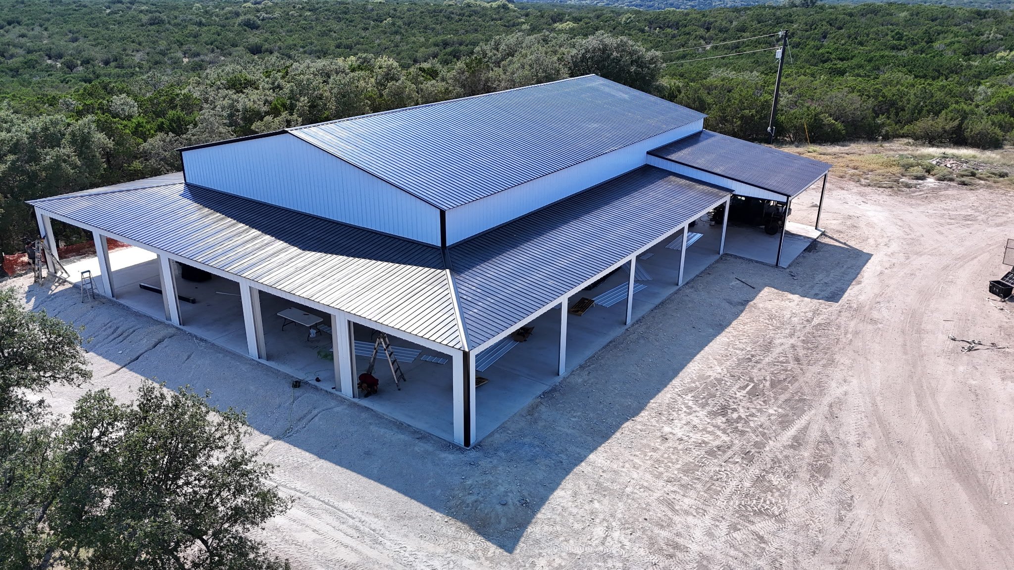 An aerial view of a large, modern, metal-roofed building under construction in a rural area surrounded by trees. The building has open sides supported by white columns, with construction materials and equipment visible around it.