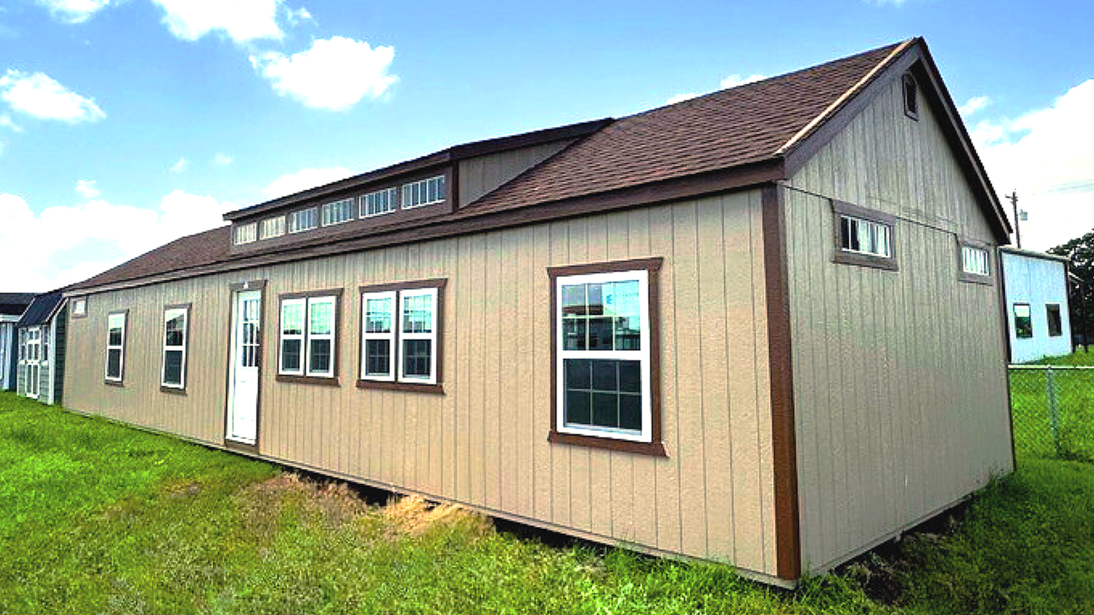 A beige manufactured home with white trim and multiple windows, situated on a grassy lot with a chain-link fence partially visible on the right side.