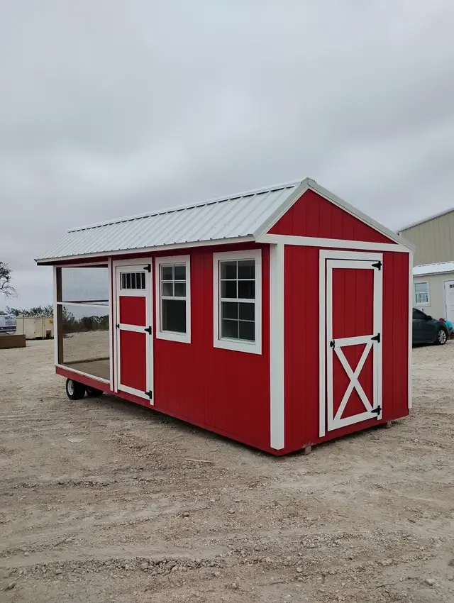 A small red barn-style chicken coop with white trim, a metal roof, and wheels, situated on a gravel lot.