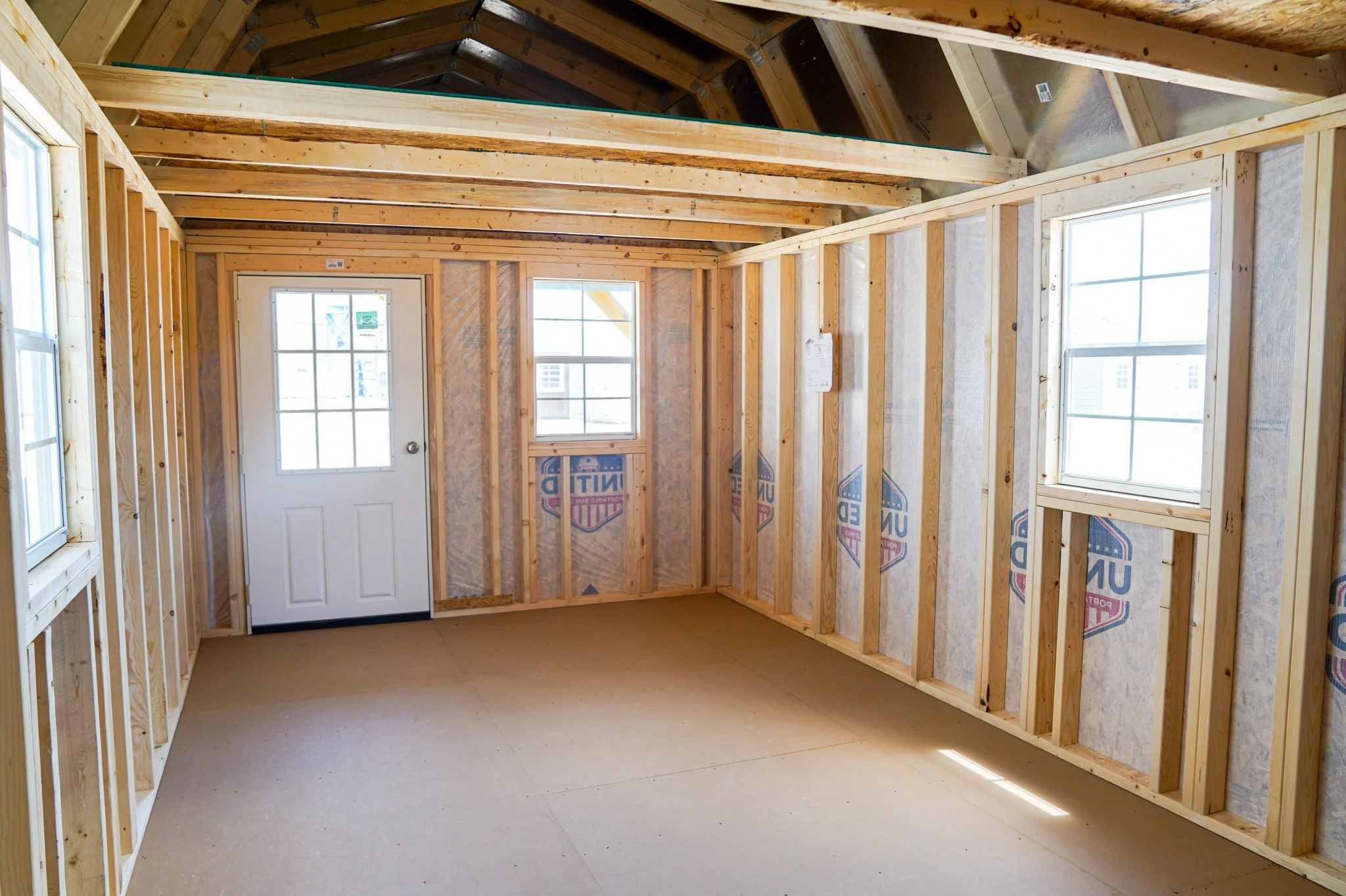 Interior view of an unfinished room in a portable building with exposed wooden framing, three windows, and a door with glass panes. The ceiling is vaulted with wooden beams.