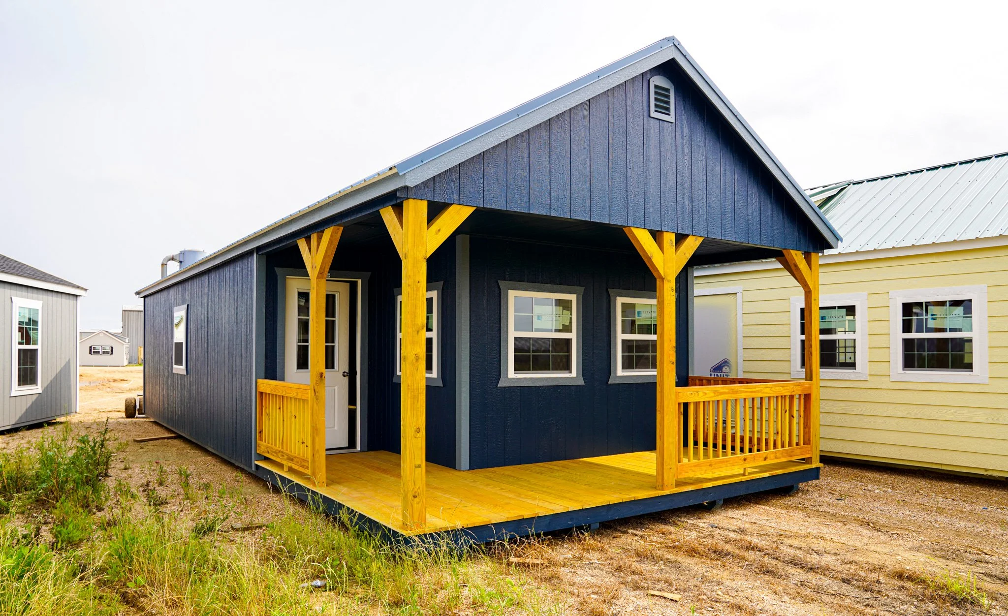 Small blue cabin with a front porch, yellow railings and supports, and a grey metal roof, surrounded by dirt and grass, with other similar houses in the background.