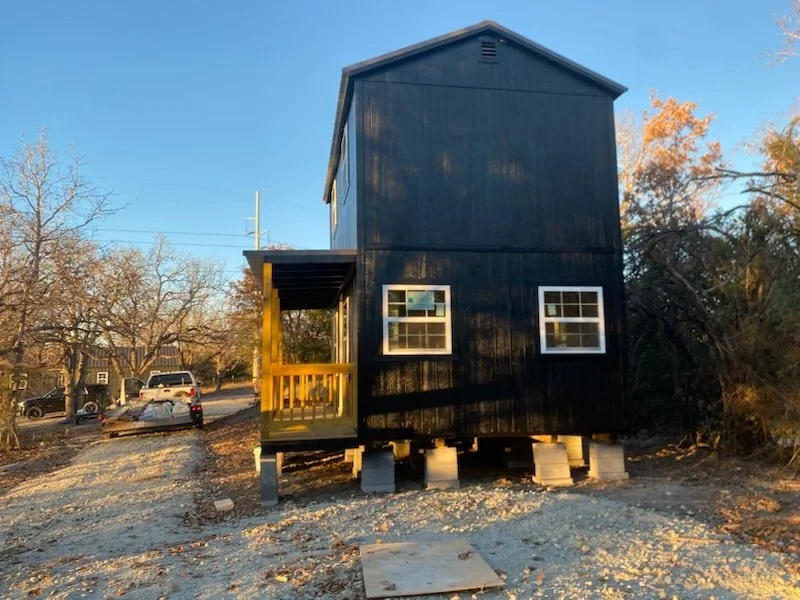 A large 2 story elevated black tiny home with a porch, two windows, and a sloped roof, built on concrete stilts in a rural area with trees and parked vehicles nearby.