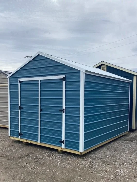 A blue metal shed with white trim and double doors, situated on a gravel lot.