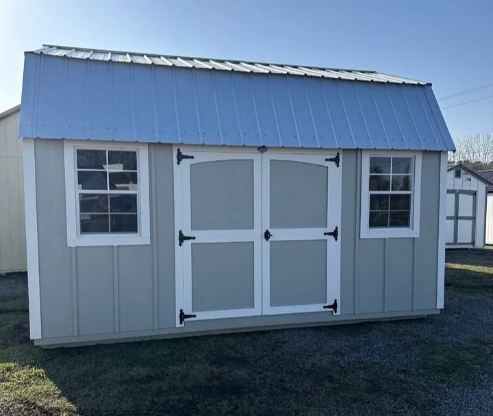 Small outdoor shed with gray siding, white trim, double doors, and two windows, on a grassy area under a clear blue sky.