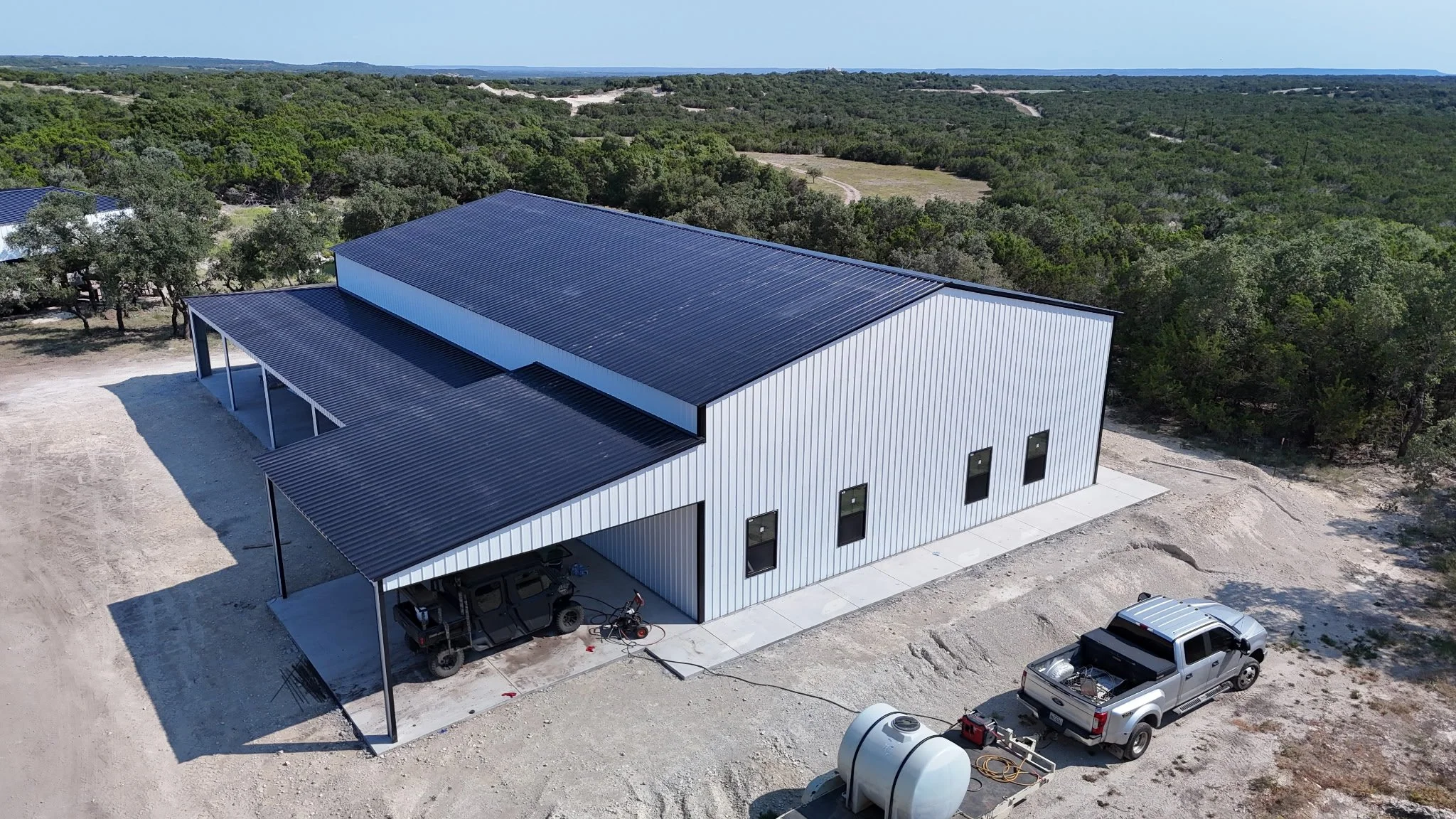 A large modern metal building with a sloped roof, situated in a rural area surrounded by trees and open land. There are vehicles parked underneath a covered area, a truck connected to a trailer, and construction equipment nearby.