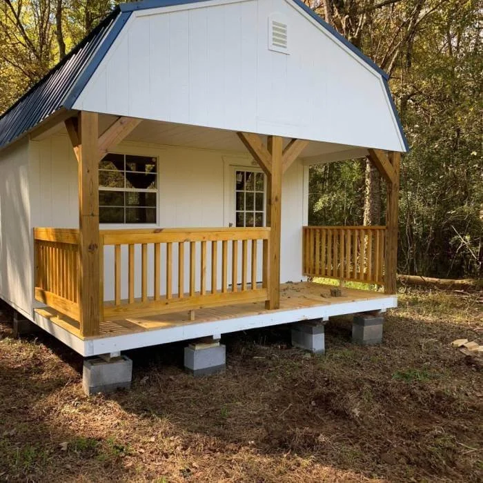 Large white cabin with a blue metal roof and a front porch with wooden railings, elevated on concrete blocks, surrounded by trees.