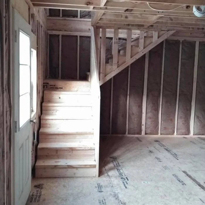 Interior of a house under construction, showing a staircase leading to a second floor, surrounded by exposed wooden framing and unfinished floors.