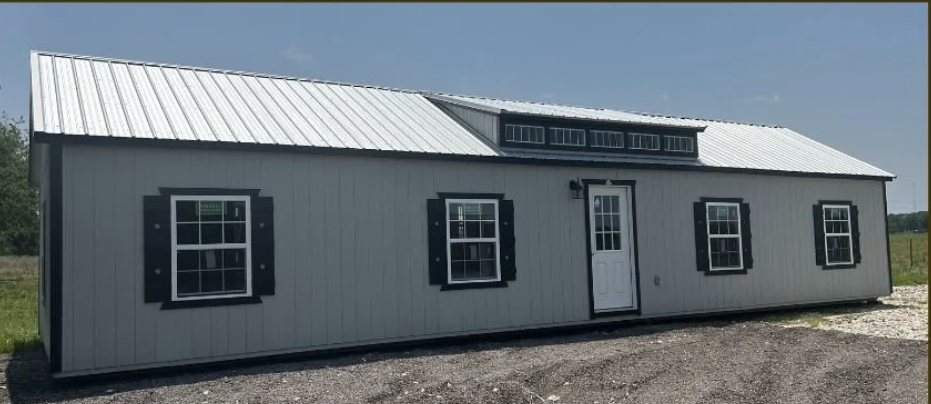 A long, white, prefabricated portable building with black shutters and a central door, set on a gravel lot under a clear blue sky.