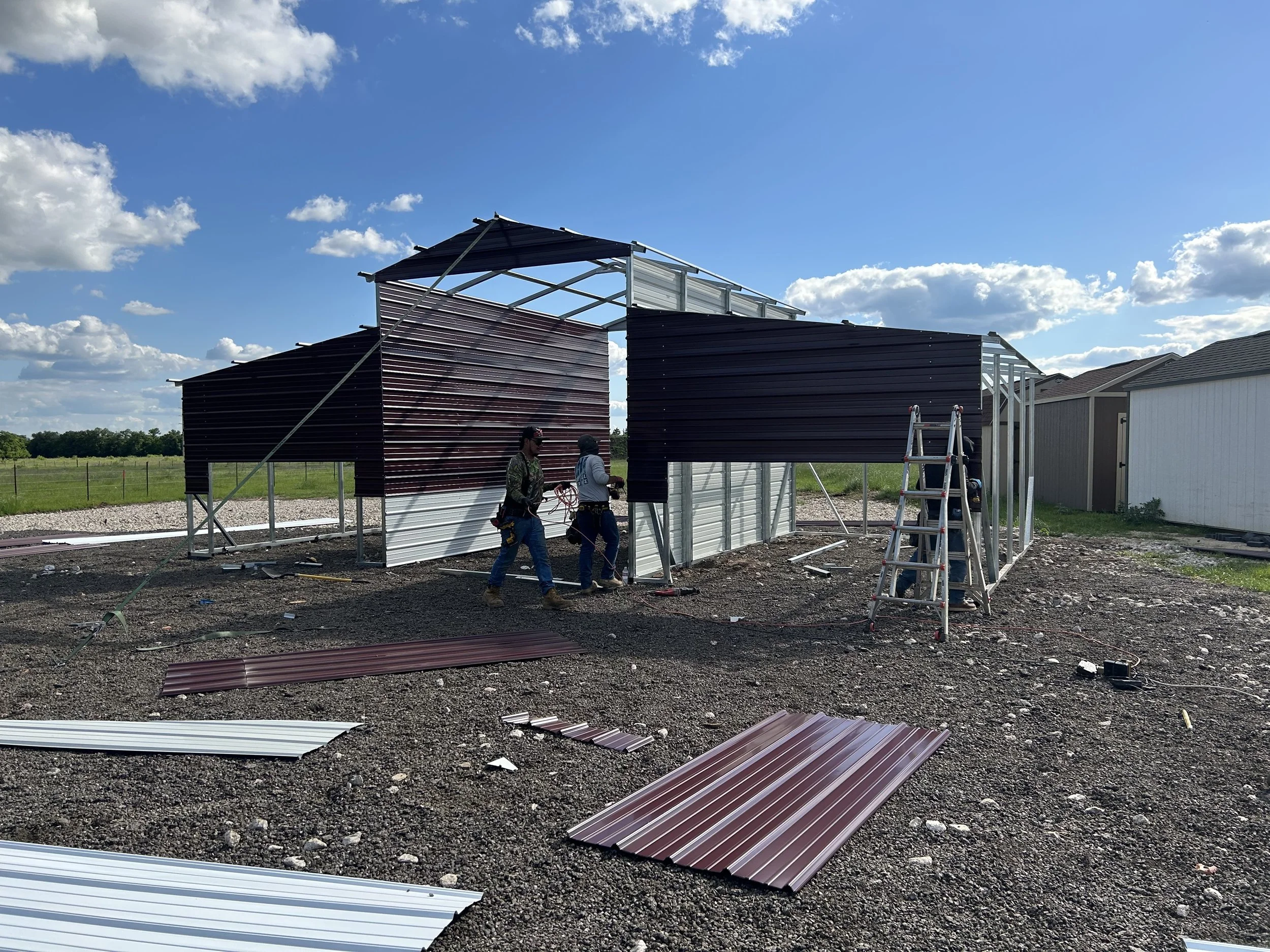 Two construction workers assembling a metal building with dark red and silver metal panels on a gravel lot under a blue sky with scattered clouds.
