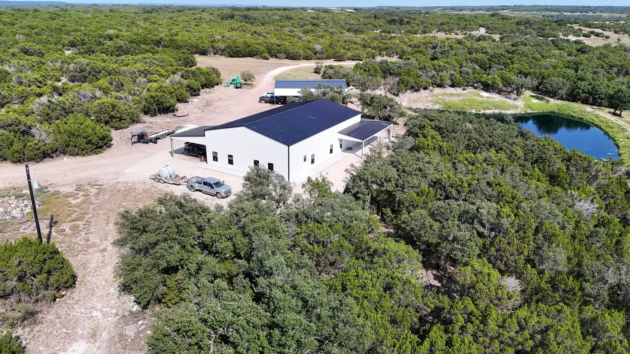 An aerial view of a modern white building with a black roof, surrounded by dense greenery and a small pond on the right side, with a dirt driveway and several vehicles parked nearby.