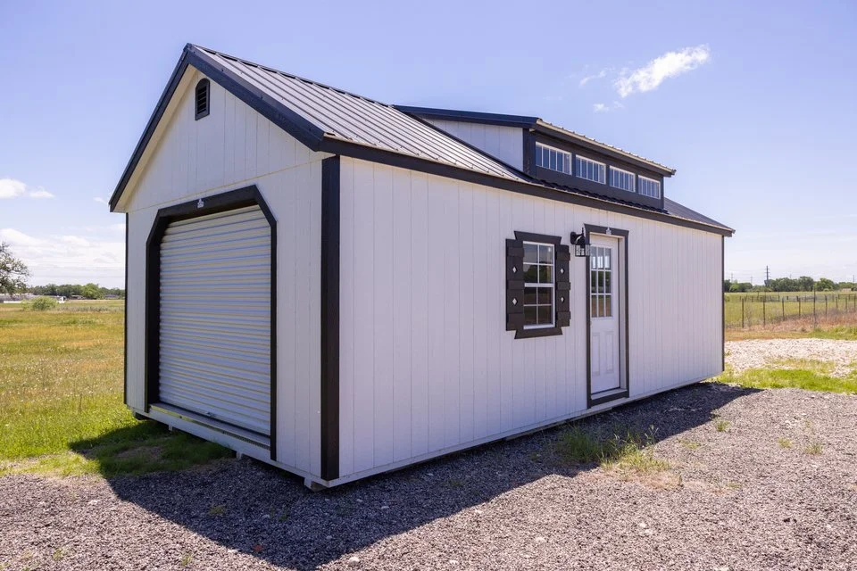 A small white and black garage shed with a metal roof in an open grassy area under a blue sky.