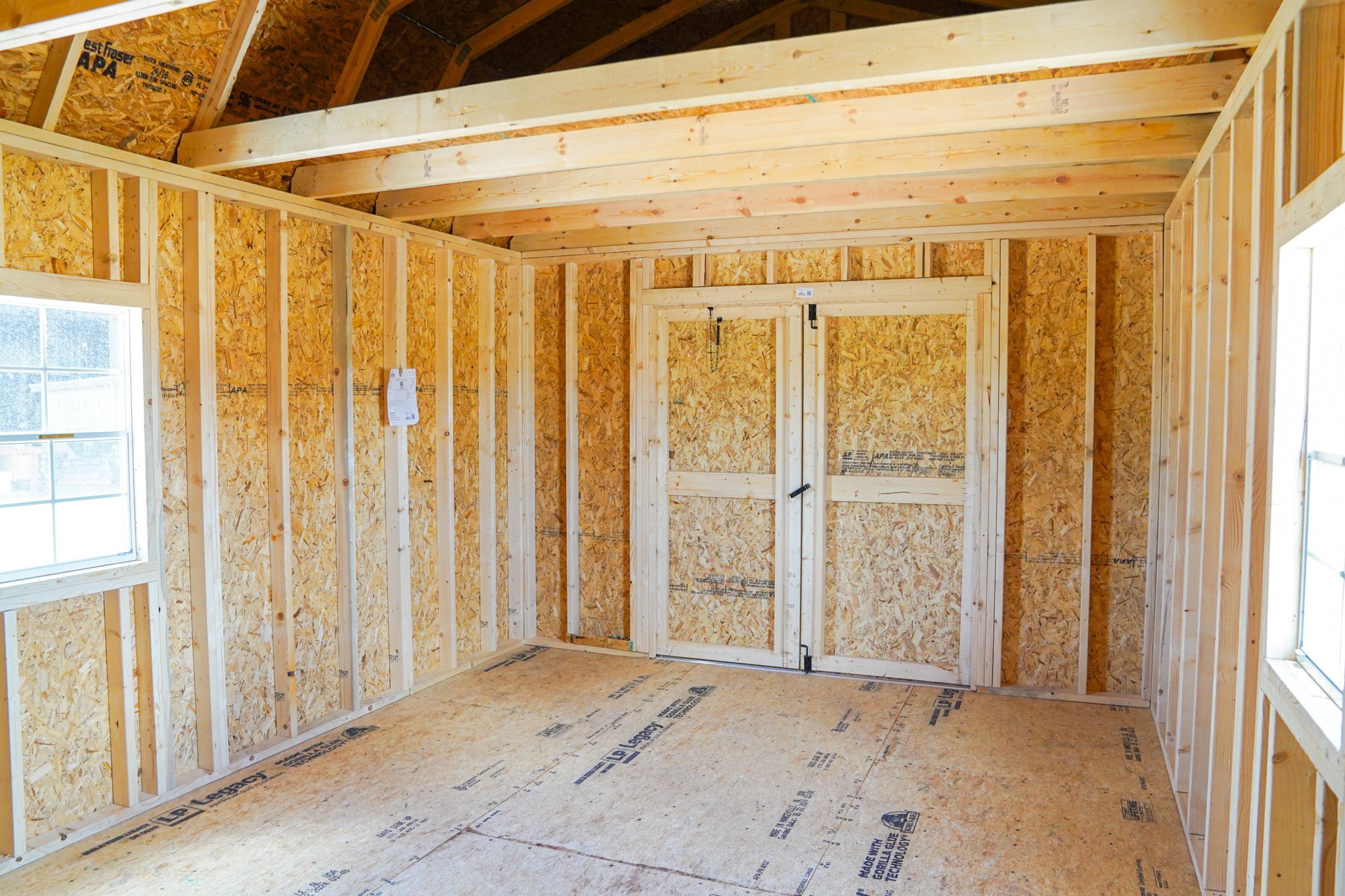 Interior of a partially constructed room with exposed wooden studs and OSB walls, including a door frame without a door and windows on two sides.