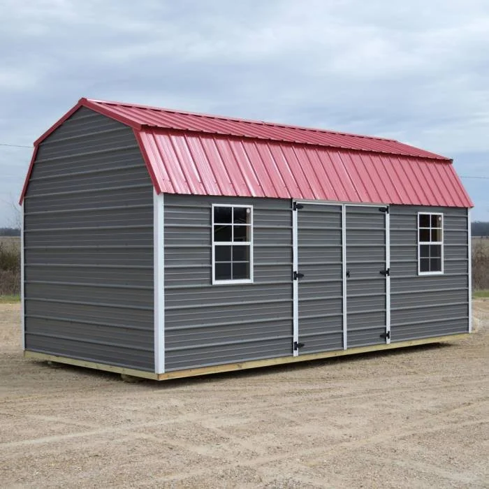 Gray metal shed with a red metal roof, two small windows, and black sliding doors, situated on a dirt lot under a cloudy sky.