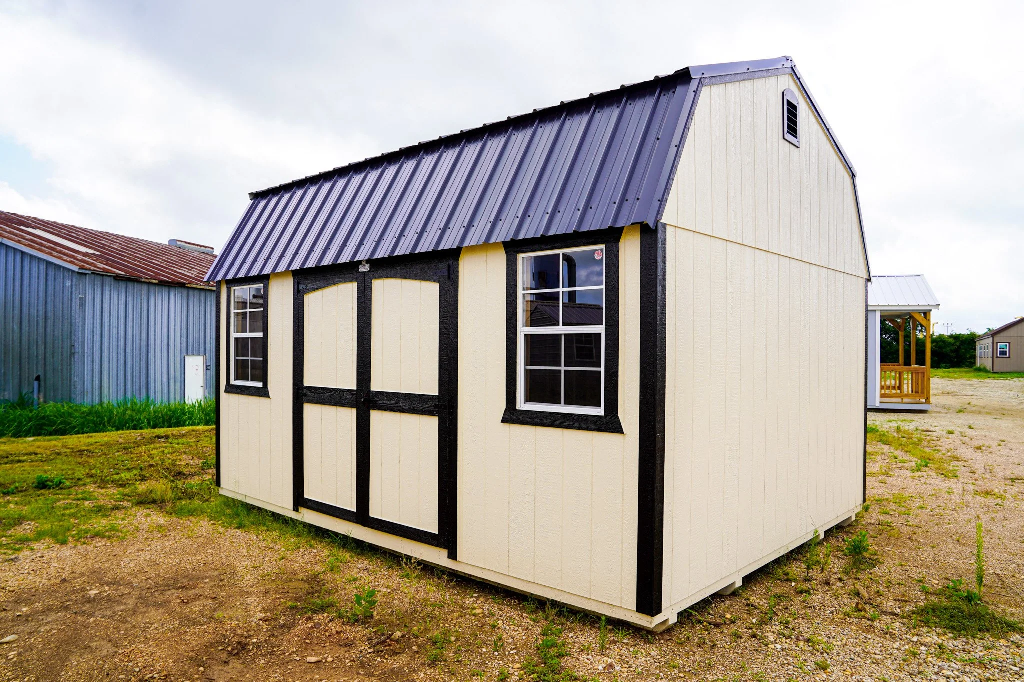 Medium sized beige shed with black trim and a metal roof, situated on a gravel lot with grass nearby, and other buildings in the background.