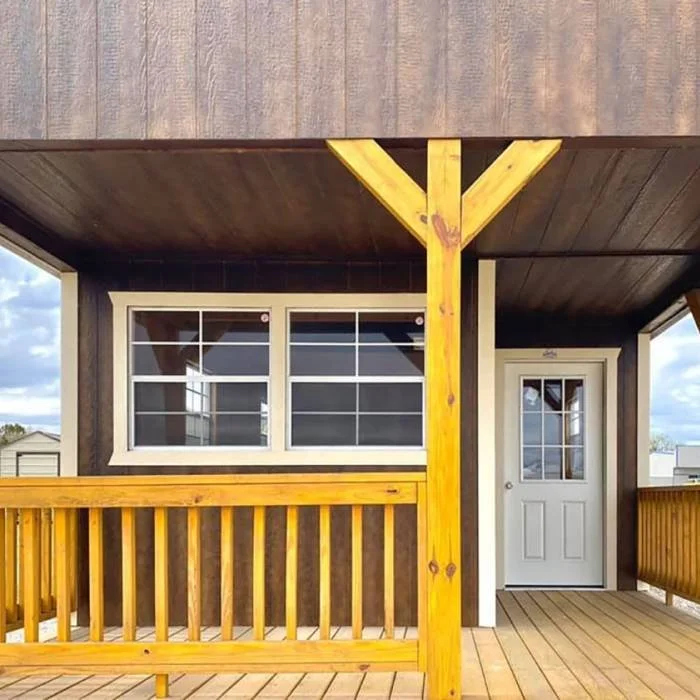 A partial view of a cabin's exterior shows a wooden porch with railings, a white door, and a window. The building has dark brown siding, and the porch ceiling and support posts are light-colored wood.