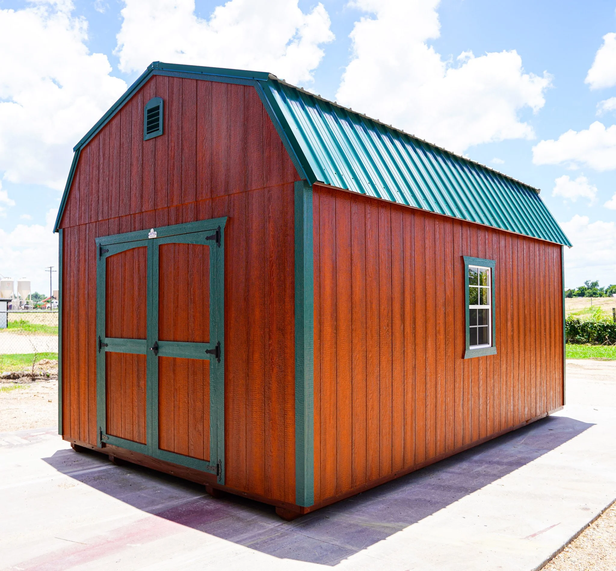A small red wooden shed with green trim and a green metal roof, featuring double doors and a window, located outdoors on a concrete surface under a partly cloudy sky.