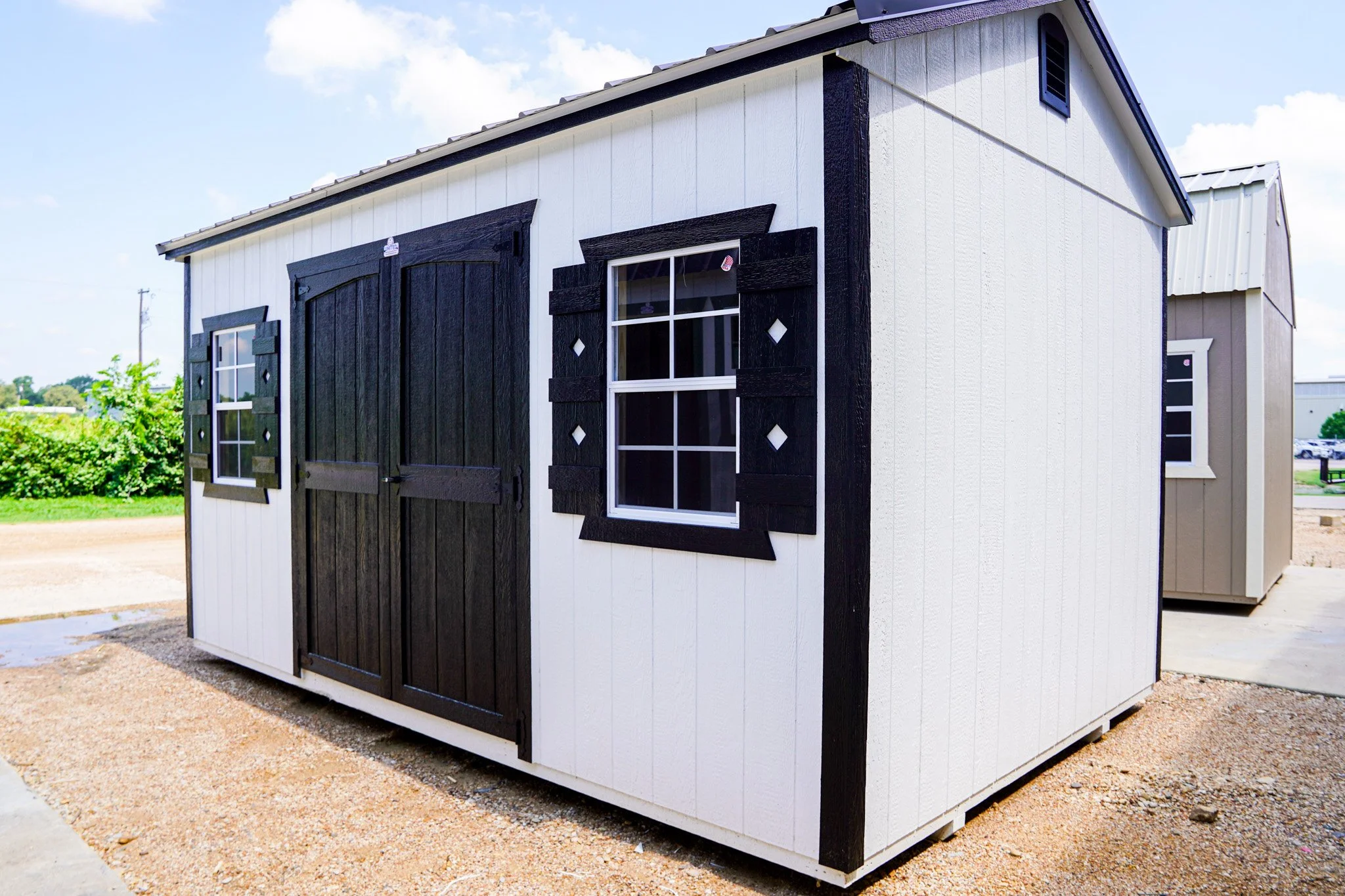 A small white shed with black trim, featuring a double door and two window shutters, situated outdoors on a gravel surface under a partly cloudy sky.