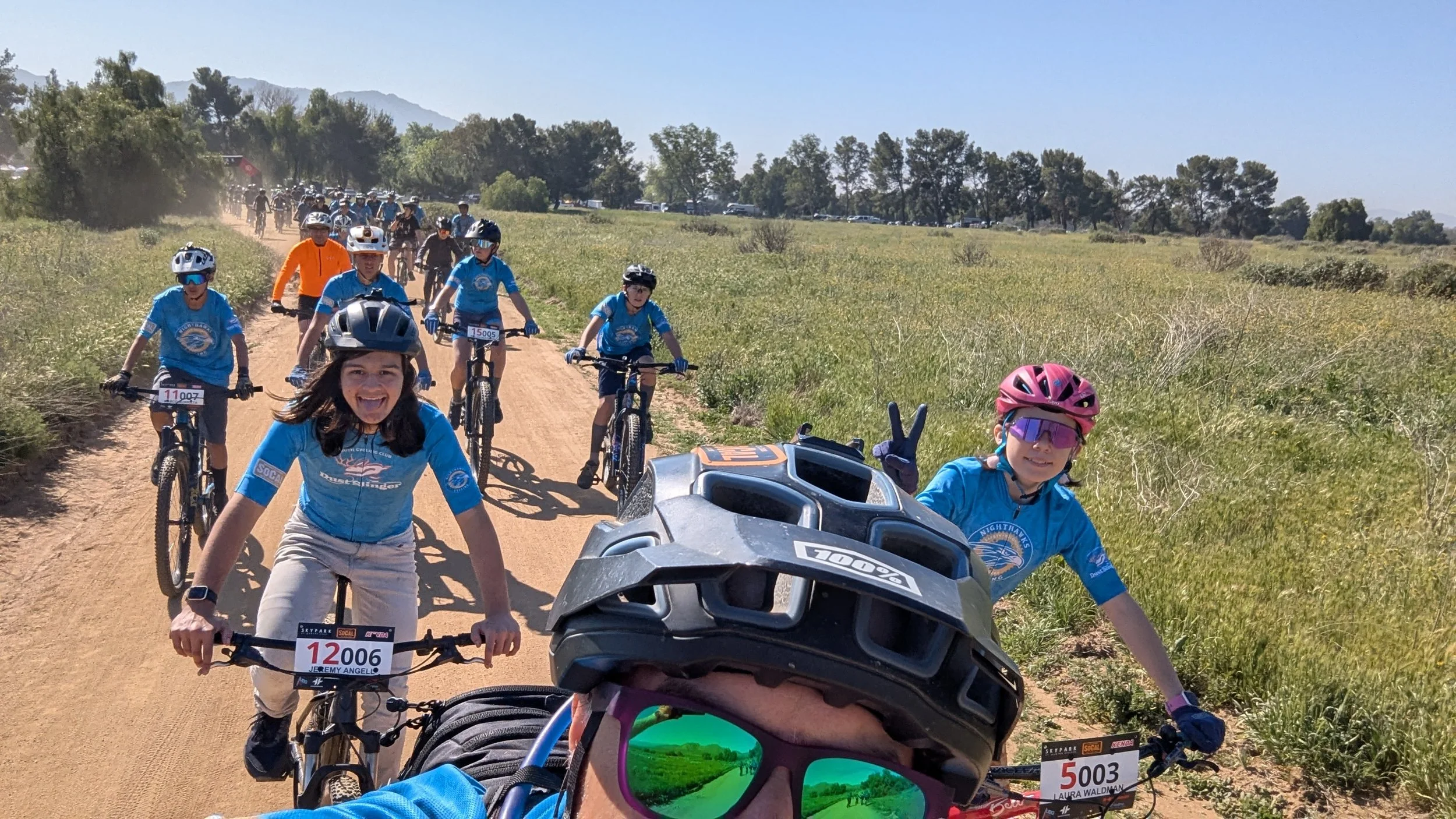 Athletes and coaches participating in a mountain biking event on a dirt trail in a hilly, rural area with trees and mountains in the background. Some children are smiling and making a peace sign, all wearing helmets and colorful cycling jerseys.