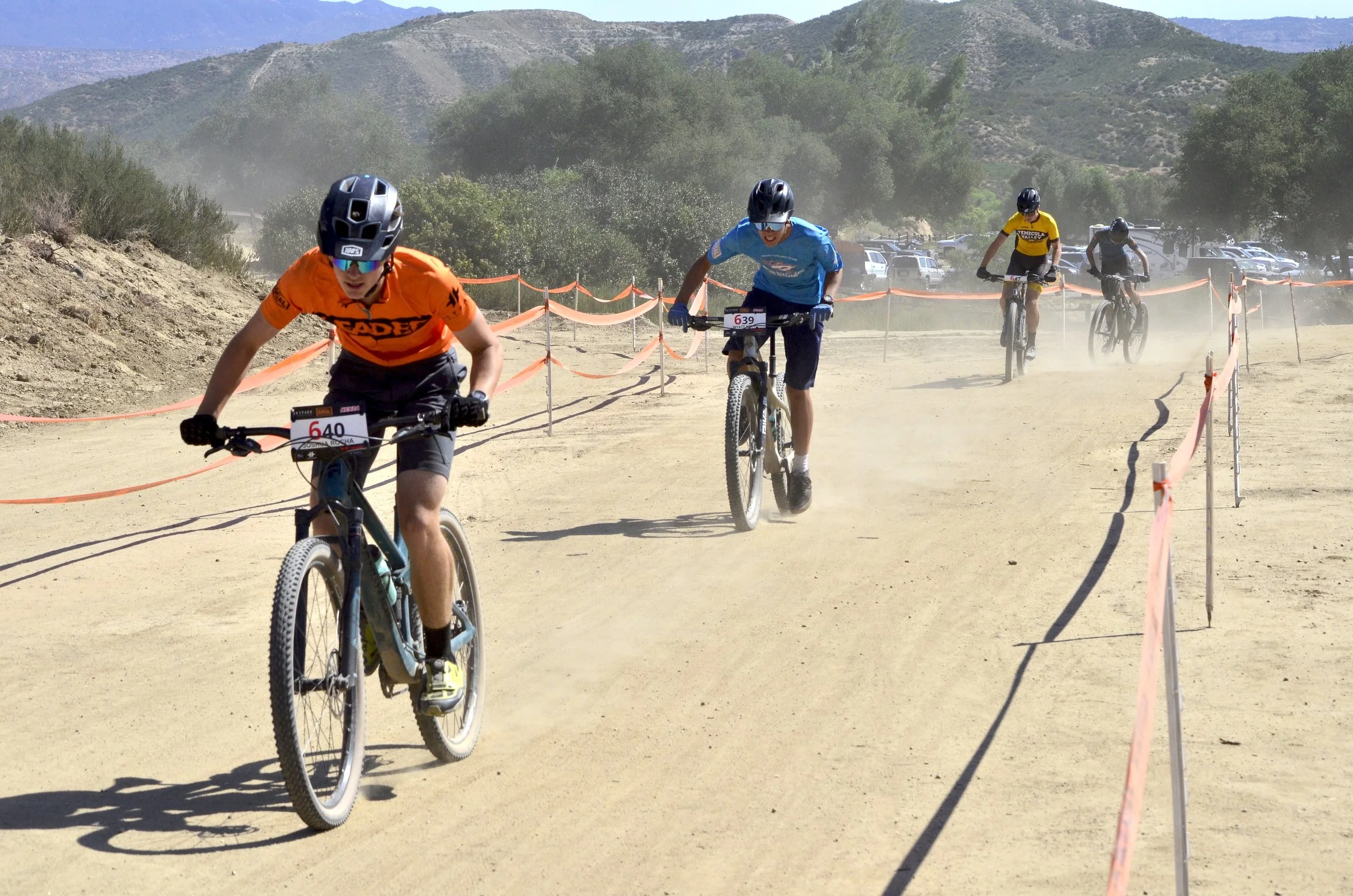 Two young boys riding mountain bikes on a dirt trail in a mountainous area with trees and distant hills, dust kicking up behind them, orange safety fence along the sides of the trail.