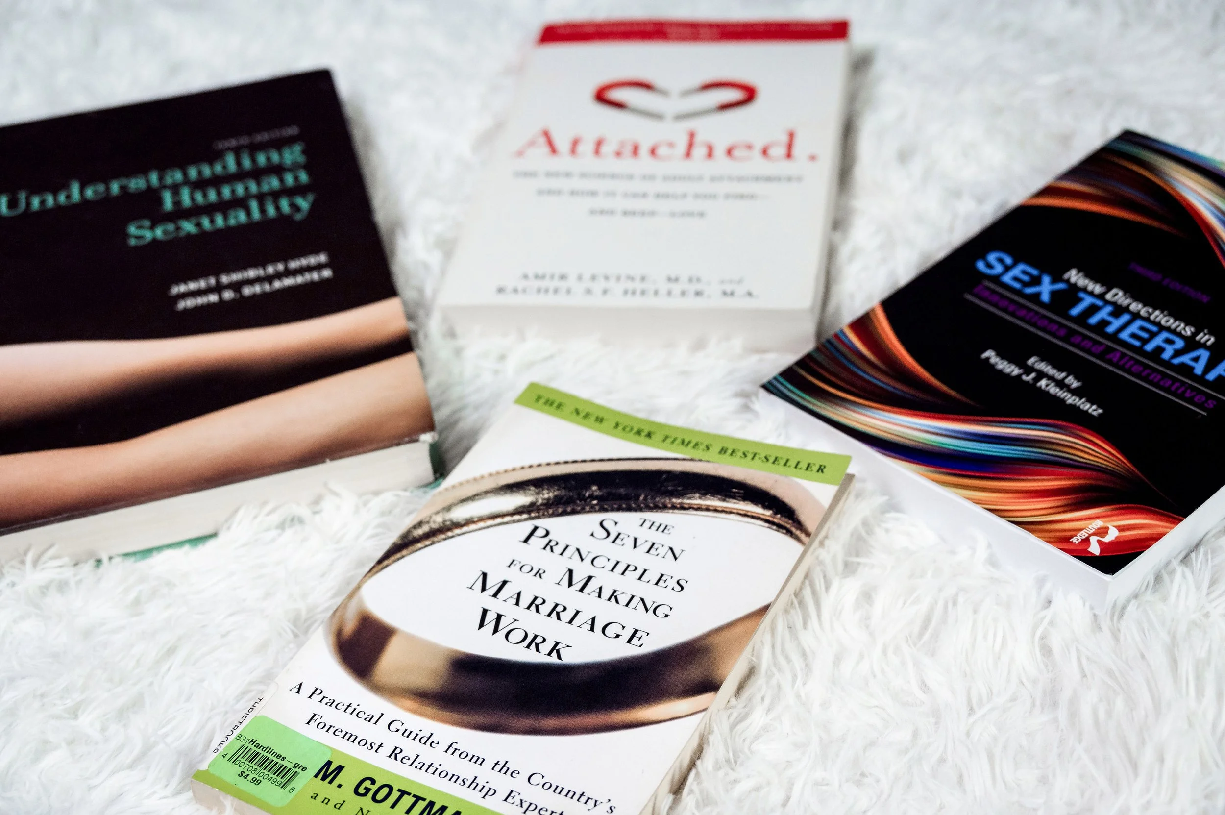 Five books placed on a white, fluffy surface, including 'Understanding Human Sexuality,' 'Attached,' 'Sex Therapy,' 'The Seven Principles for Making Marriage Work,' and a book by M. Gottman.