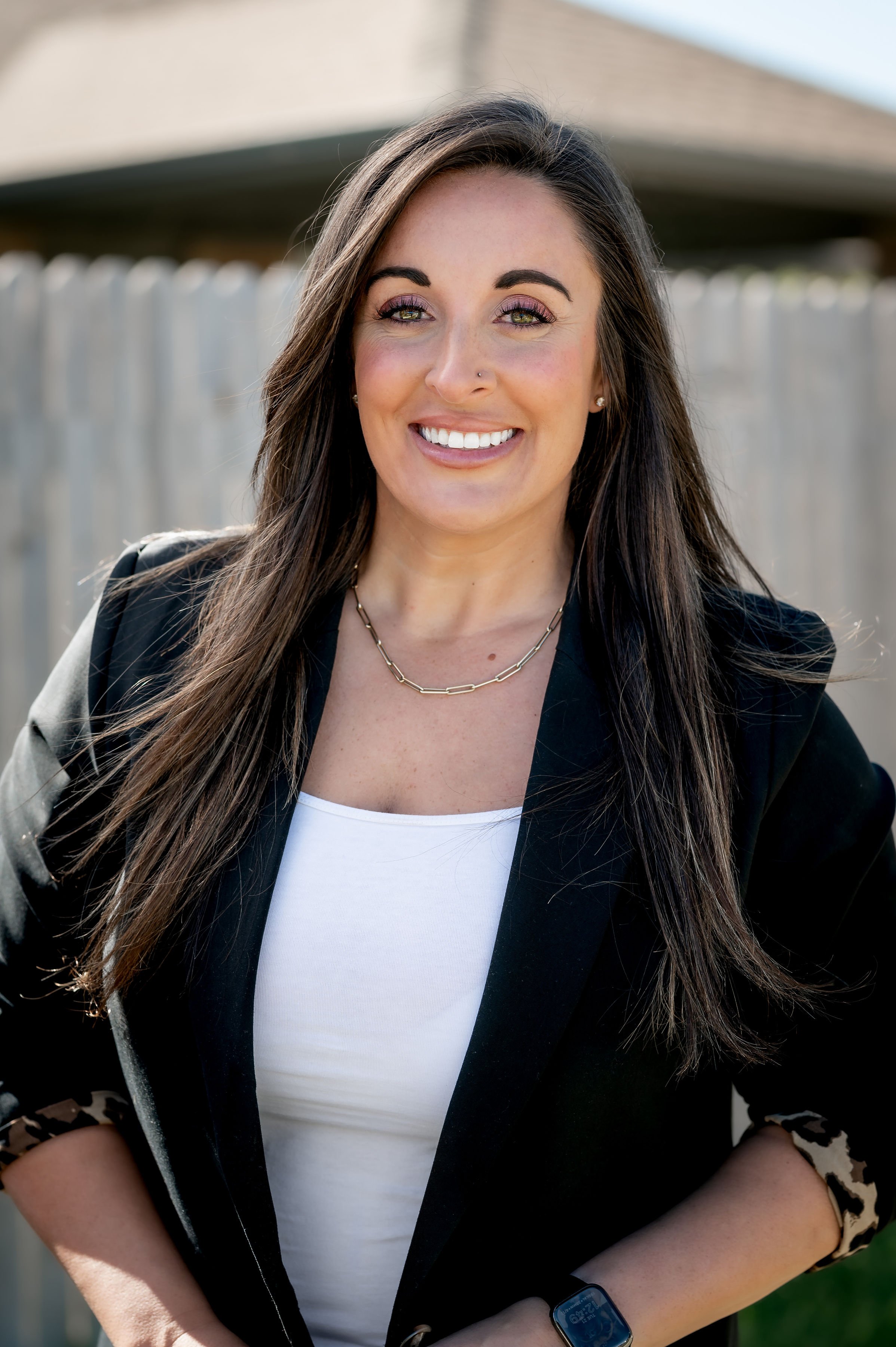 A woman with long dark hair smiling outdoors, wearing a black blazer over a white top, and a gold chain necklace.