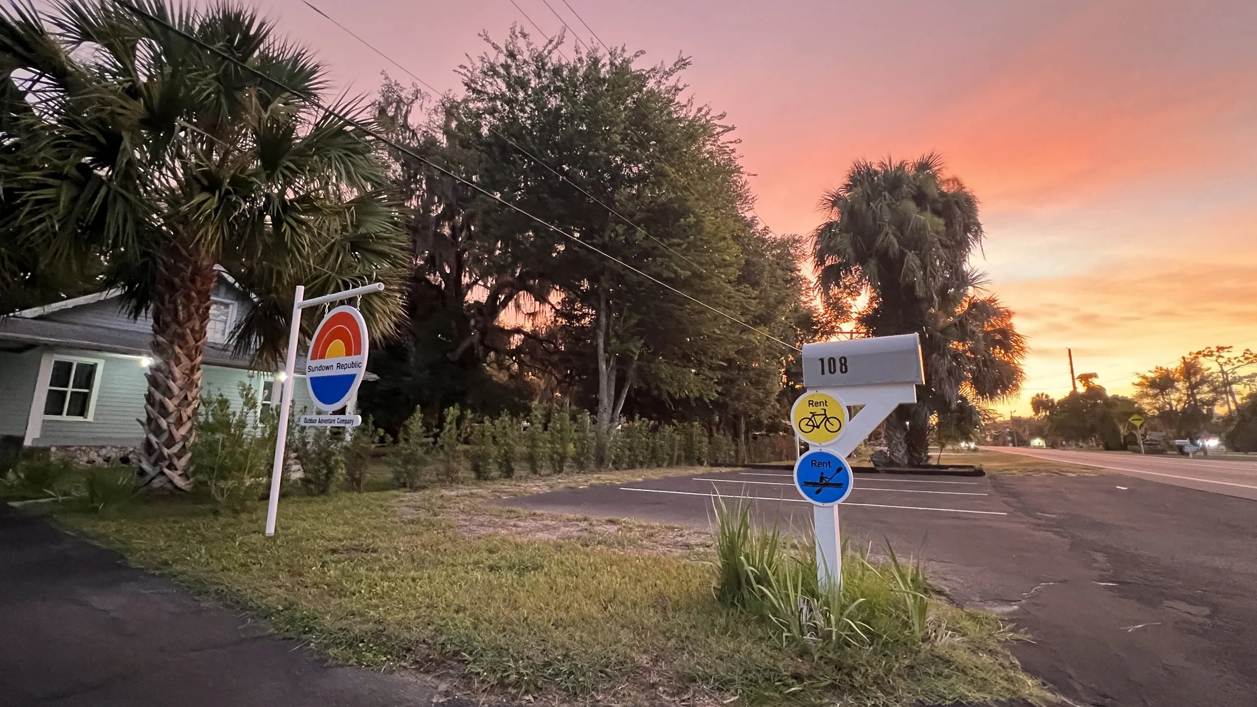 Sign reading Sundown Republic Outdoor Adventure Company and mailbox with kayak and bike rental signs with pink and orange sunset sky