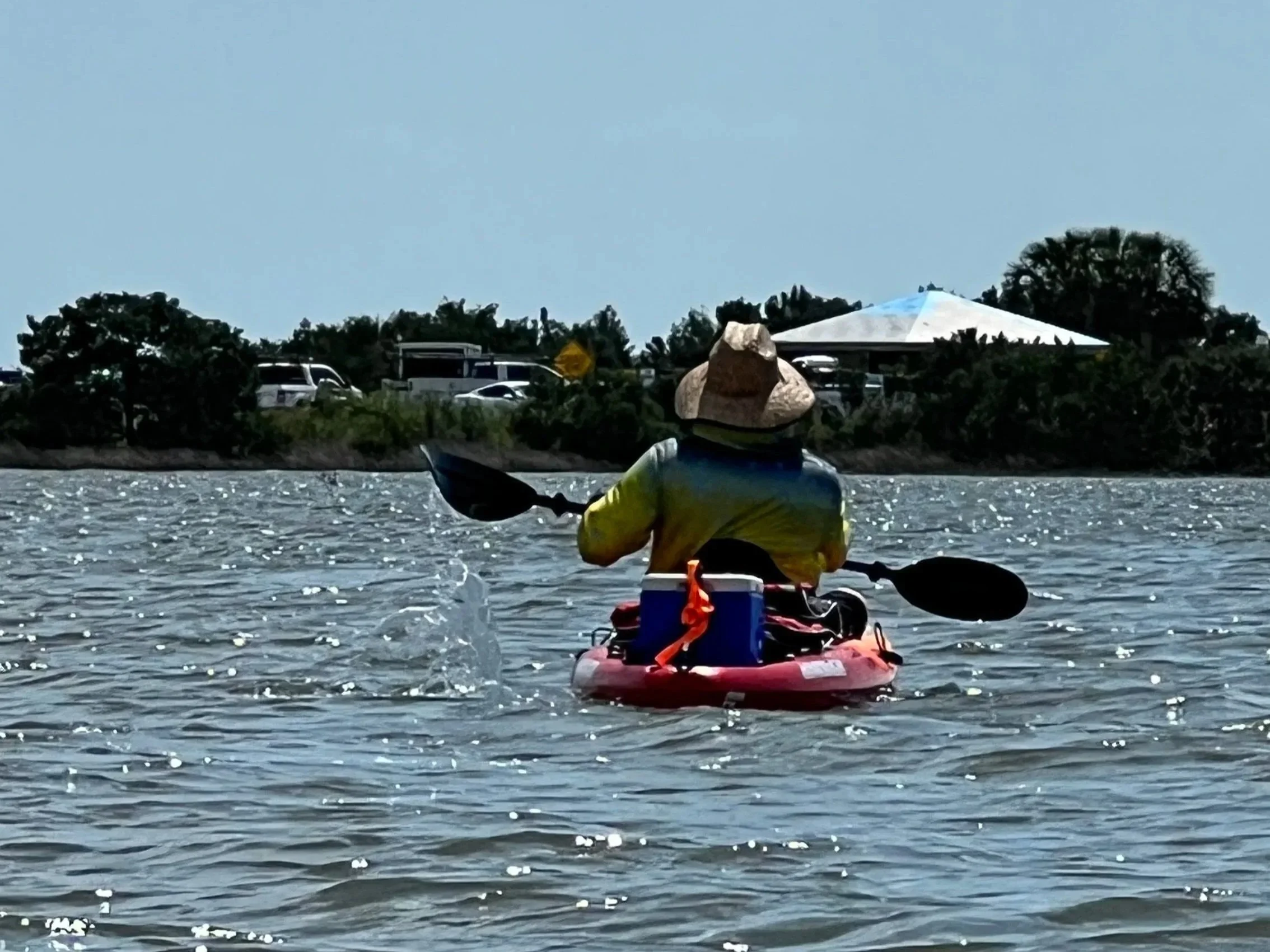 Person kayaking on water, wearing a hat, with cars and trees visible on shoreline in the background.