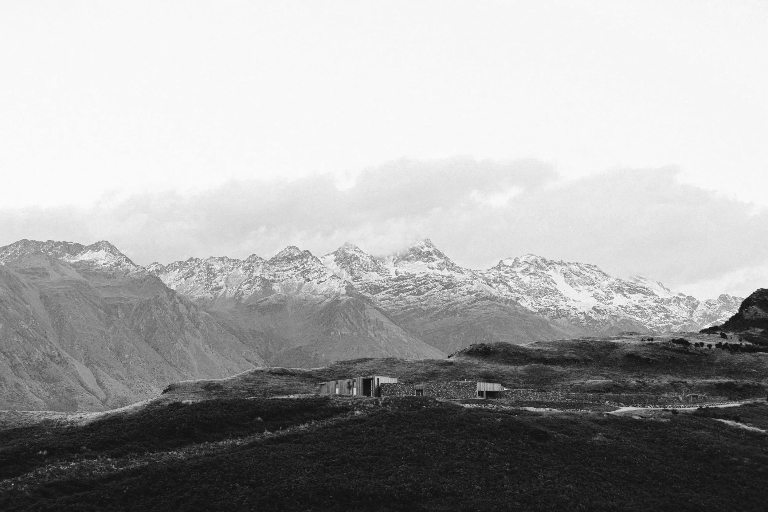 Black and white landscape of mountains with snow-capped peaks and a few modern houses on hilly terrain in the foreground.