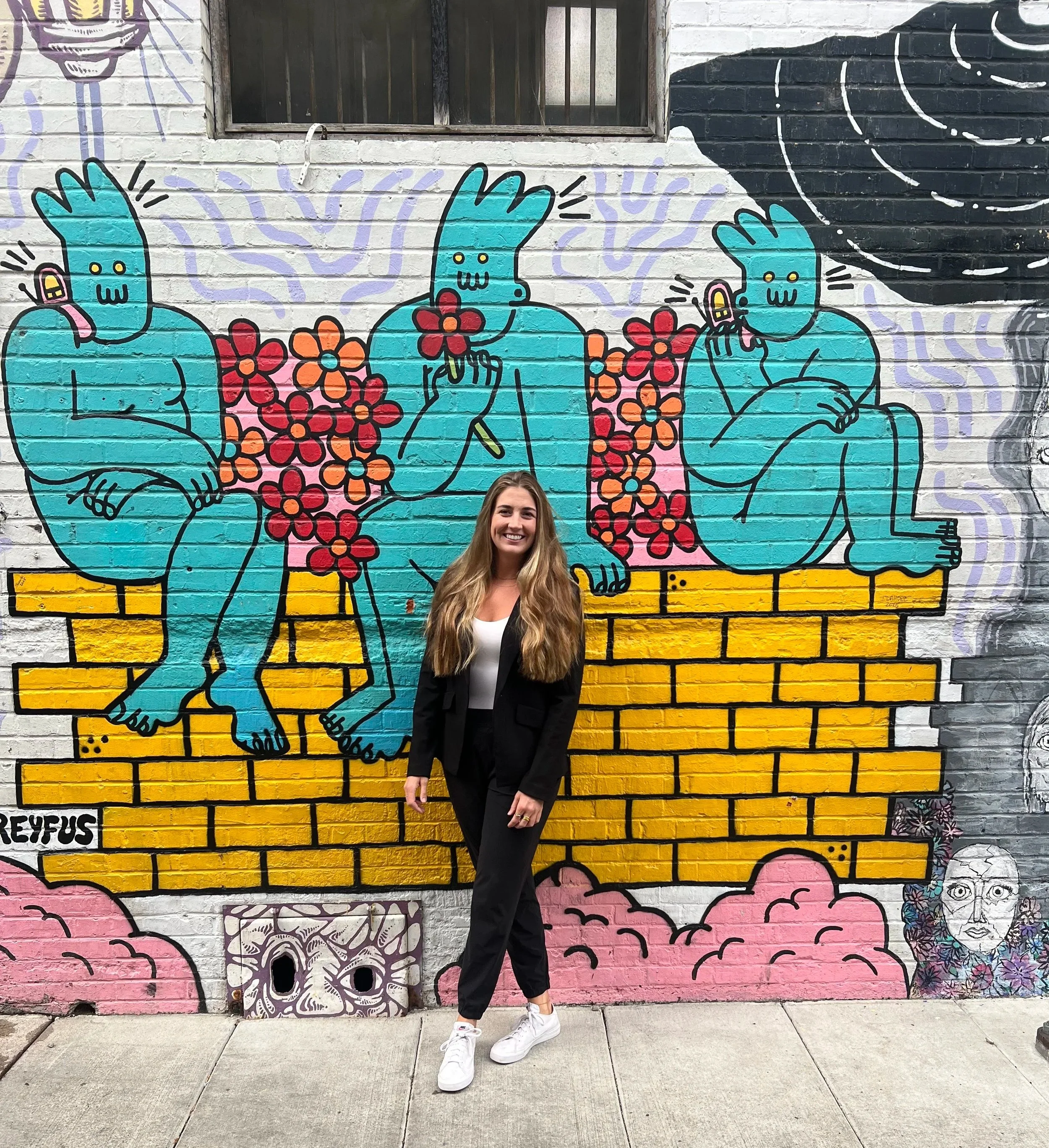 Buzz & Bloom Marketing Founder and Owner, Kaileen Koch, standing in front of a colorful mural of three blue cartoon-like figures with yellow pants sitting on a yellow brick wall, surrounded by pink and red flowers.