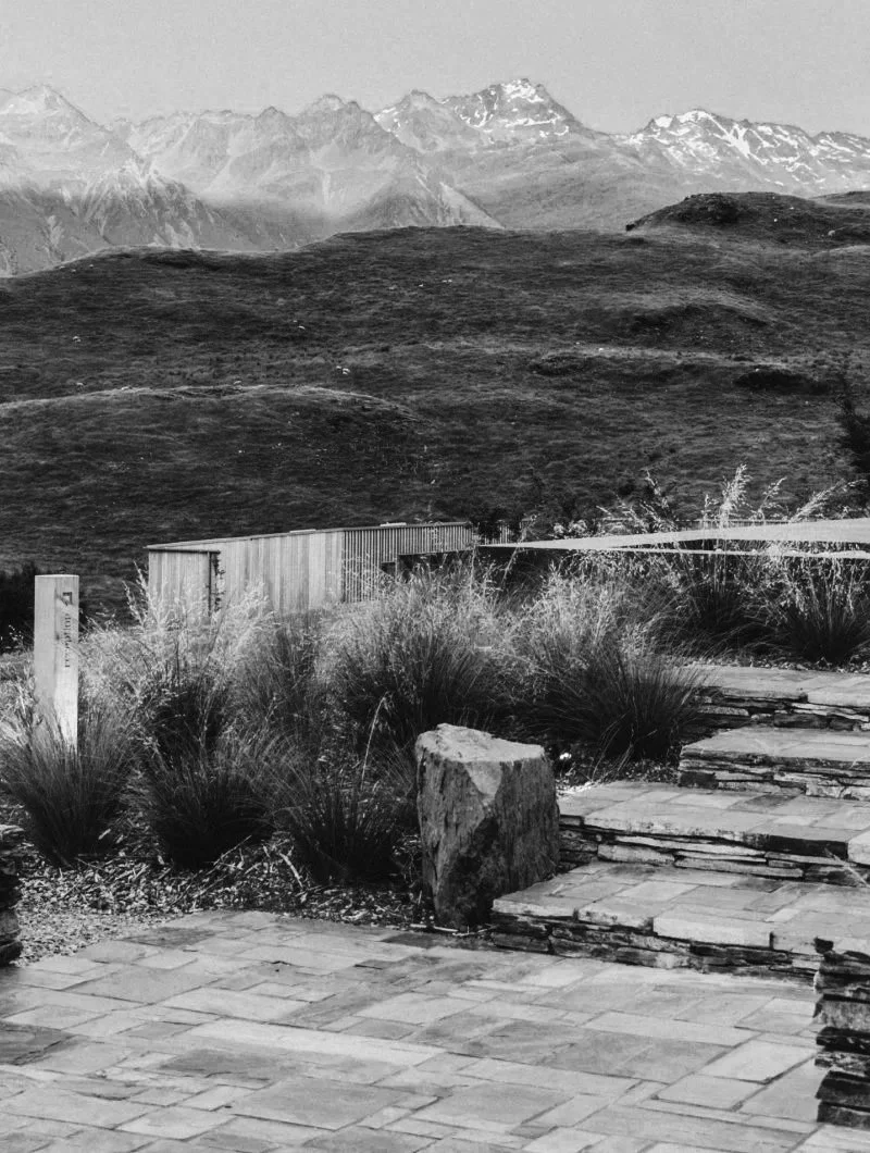 Black and white photo of a landscape with mountains in the background, hills, a stone patio with plants and rocks in the foreground, and a wooden fence and cabin in the middle ground.