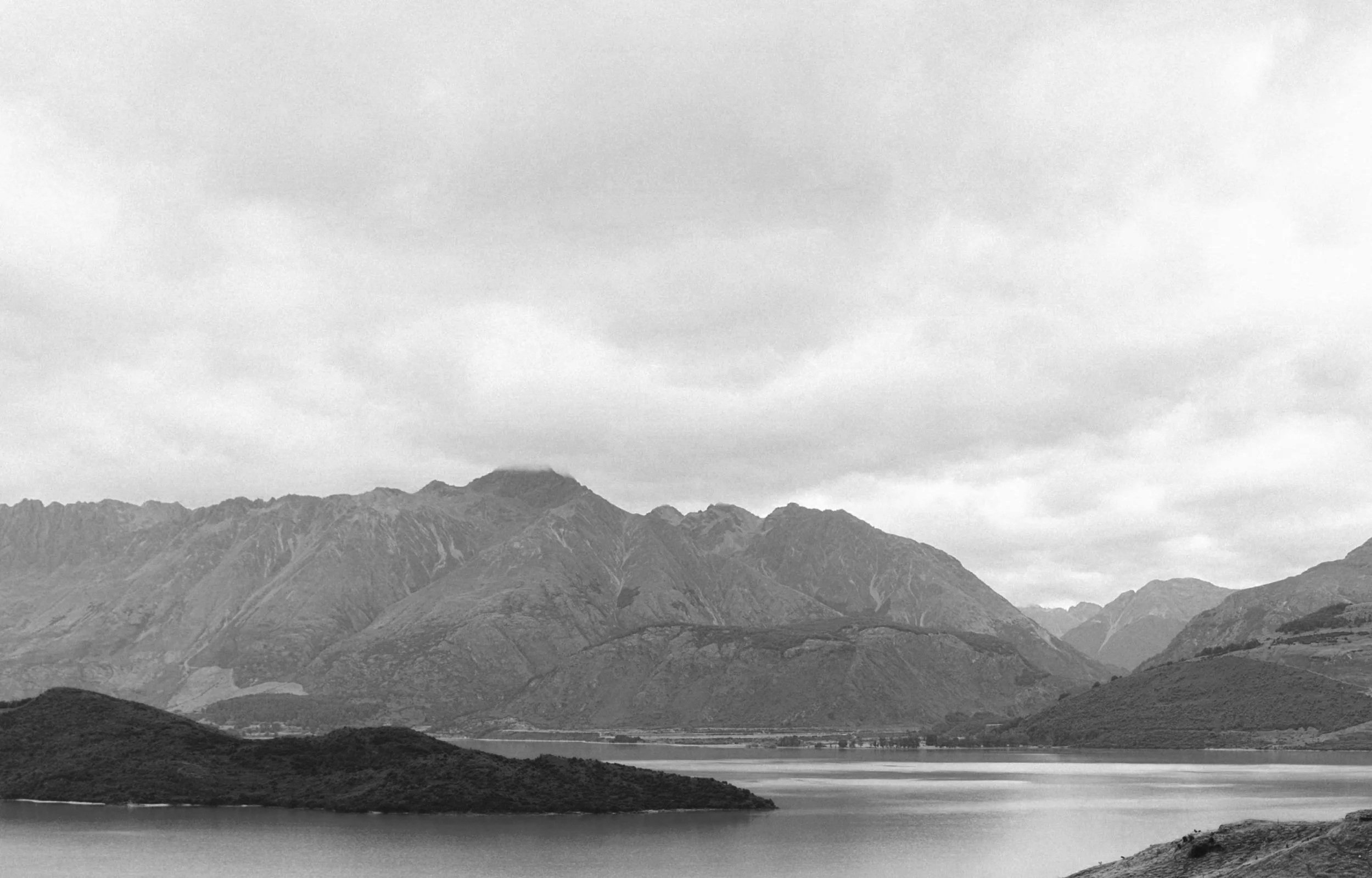 Black and white photo of mountain range with a lake in the foreground and cloudy sky above.