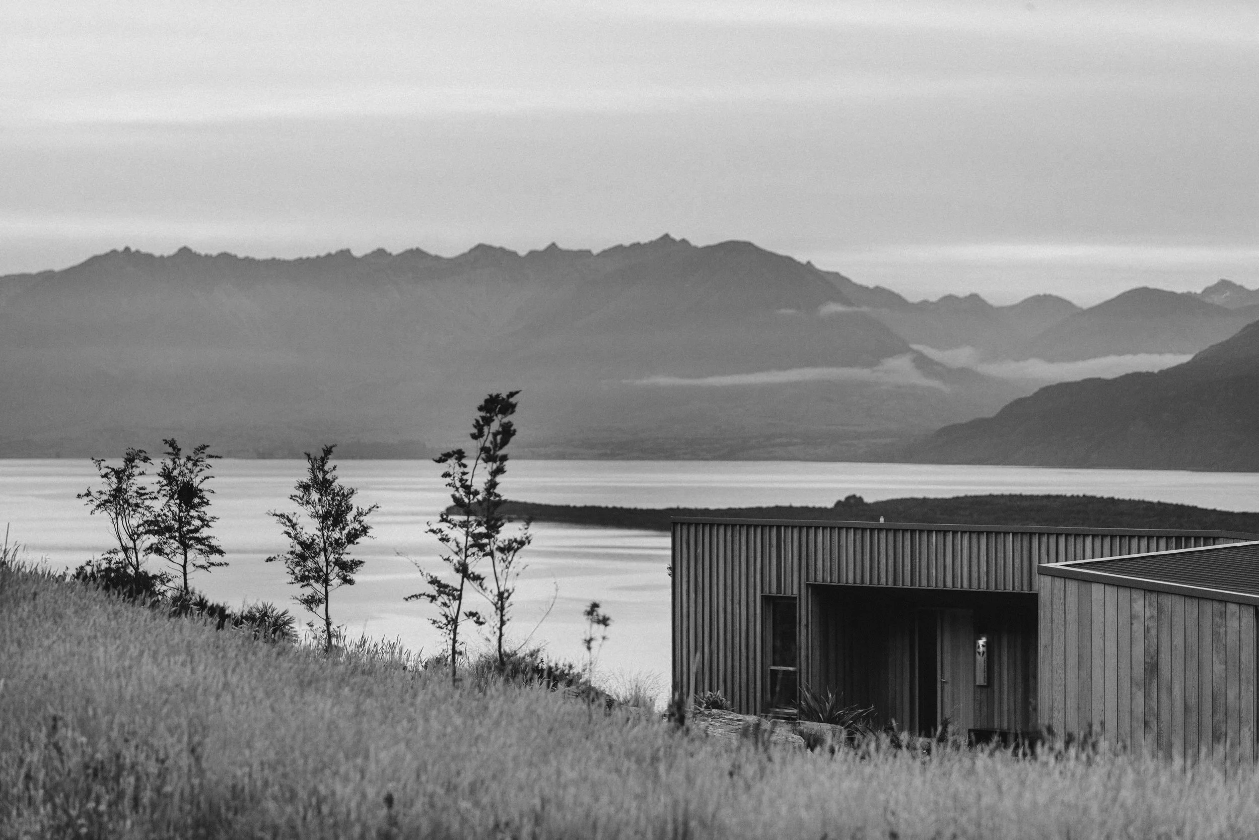 A black and white landscape featuring a modern wooden building near a body of water, with trees in the foreground and mountains in the background.