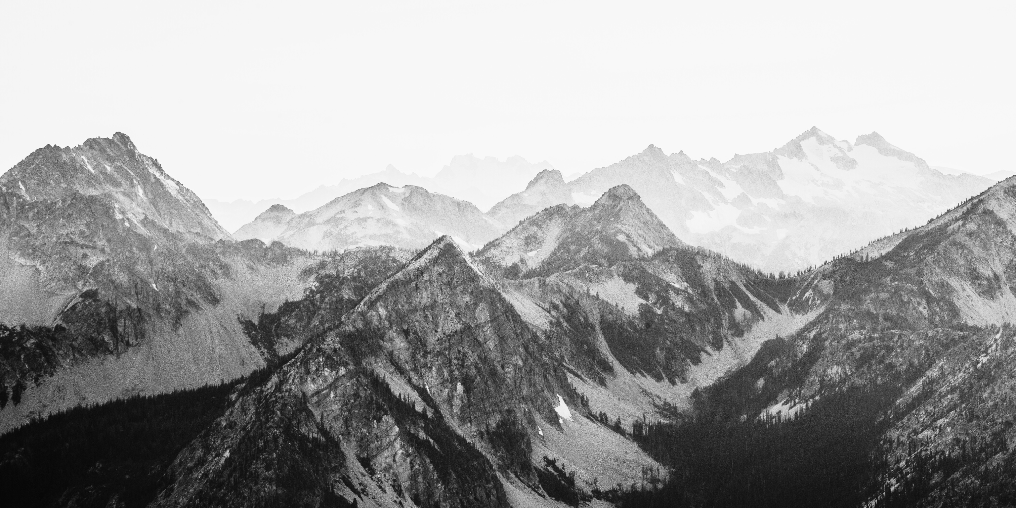 Black and white photo of rugged mountain range with snow patches, steep slopes, and dense forested areas.