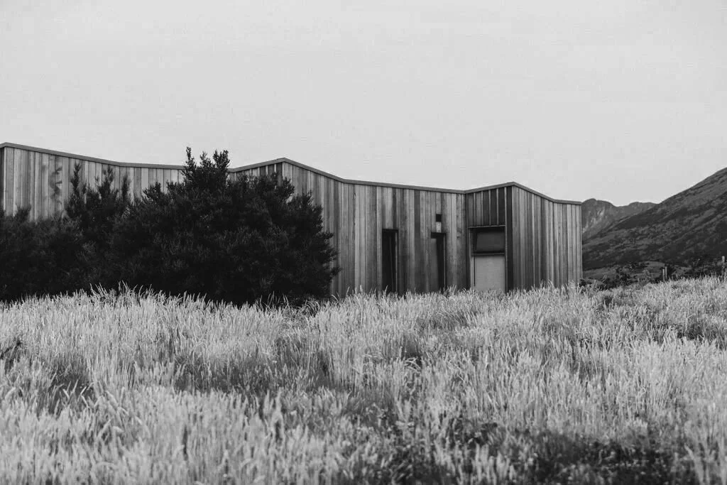 A modern, angular building with wooden exterior walls situated in a natural landscape with tall grass and mountains in the background.