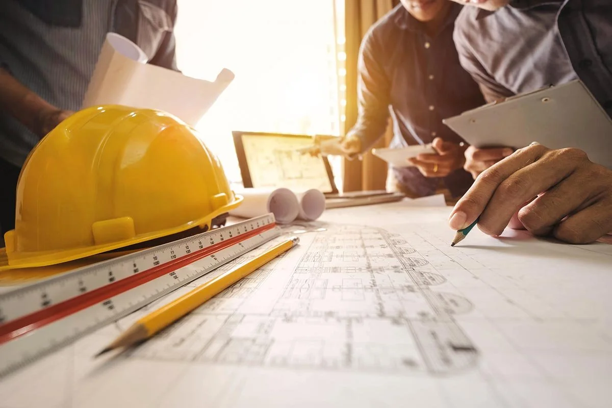 People working on construction blueprints on a table. Hard hat, ruler, and pencil in the foreground.