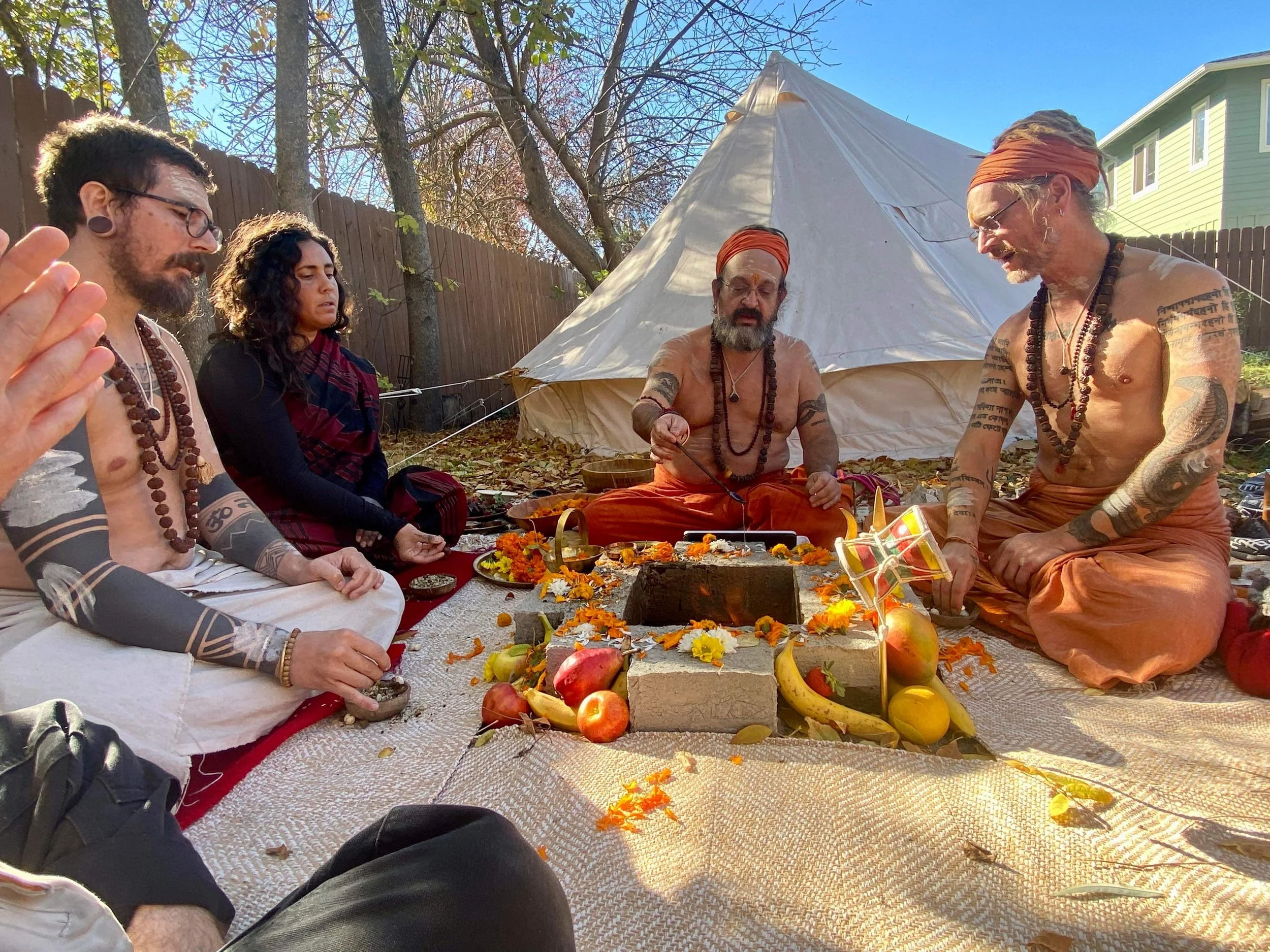 Group of people, some shirtless and with tattoos, sitting on a mat outdoors in front of a tipi. They appear to be participating in a spiritual or ceremonial gathering, with flowers and fruits on the ground.