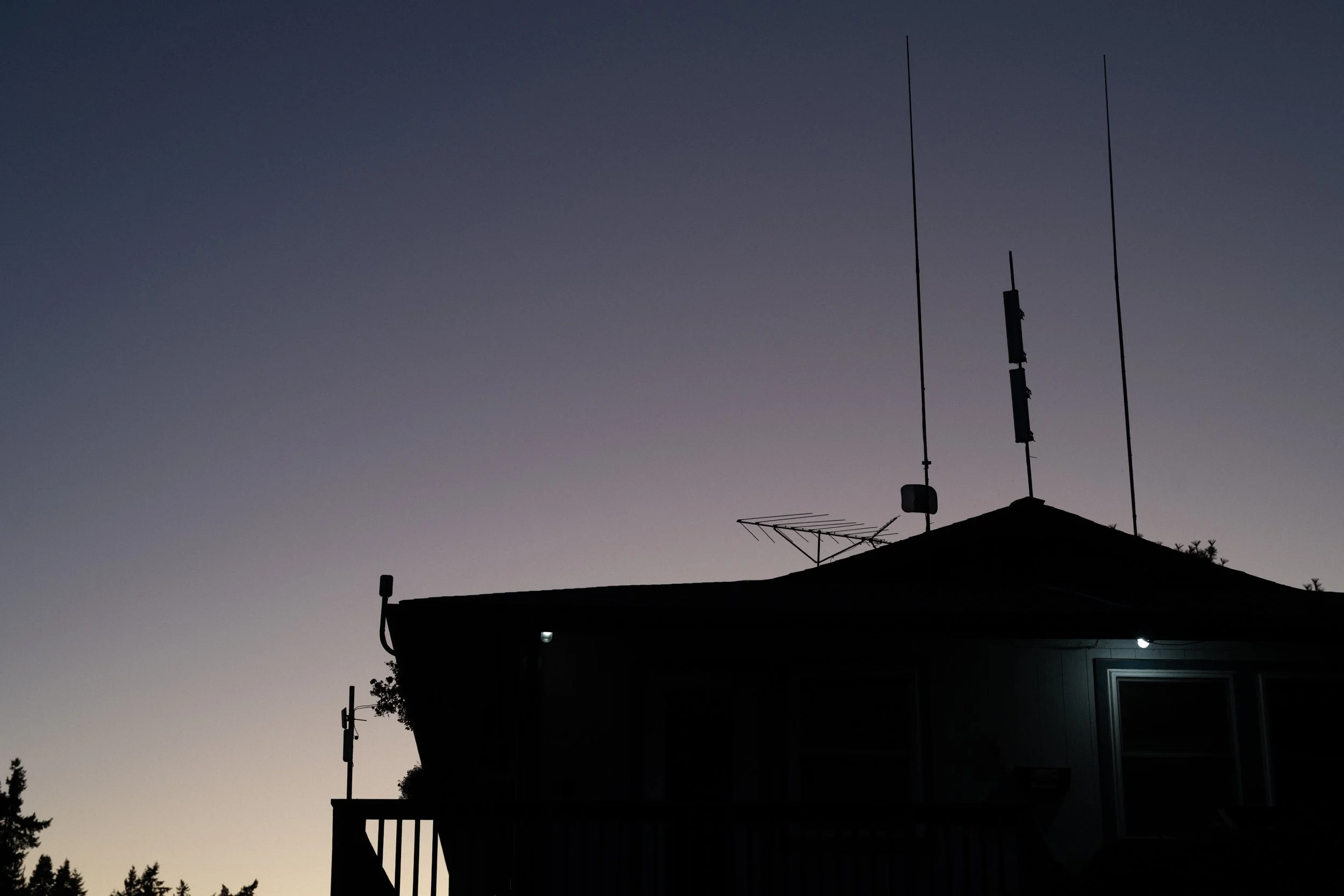 Silhouetted house roof with multiple antennas against twilight sky, some lights on in the house.