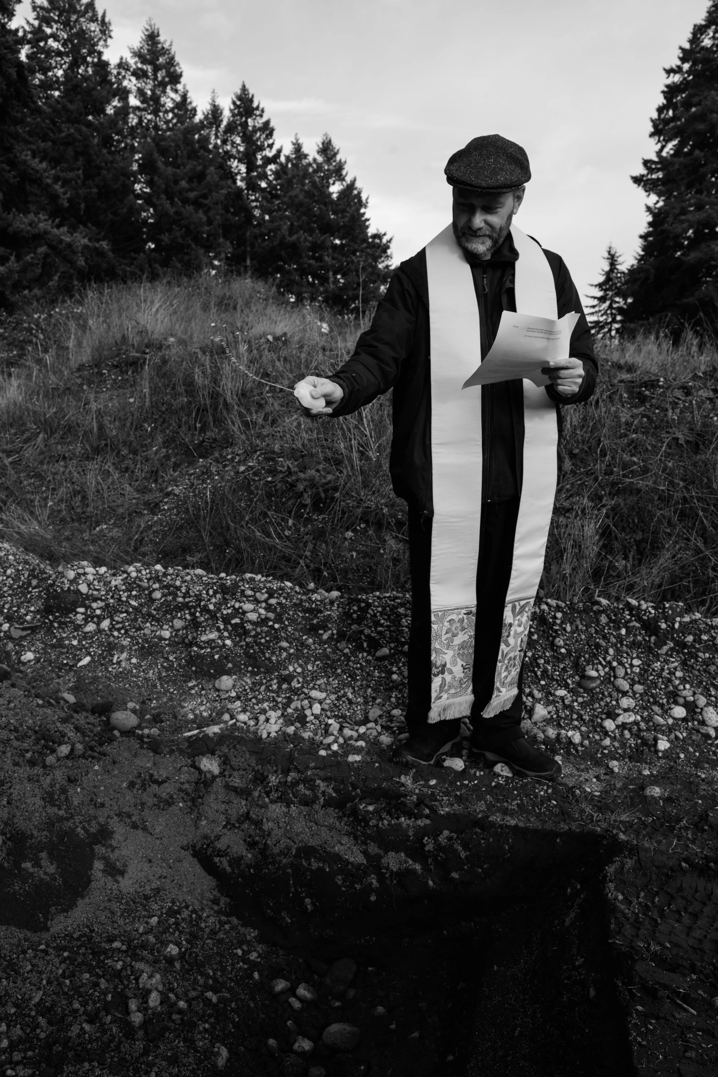 A man in religious attire, including a stole, standing outdoors near a hole in the ground, holding a document in one hand and a small object in the other, with trees and grass in the background.