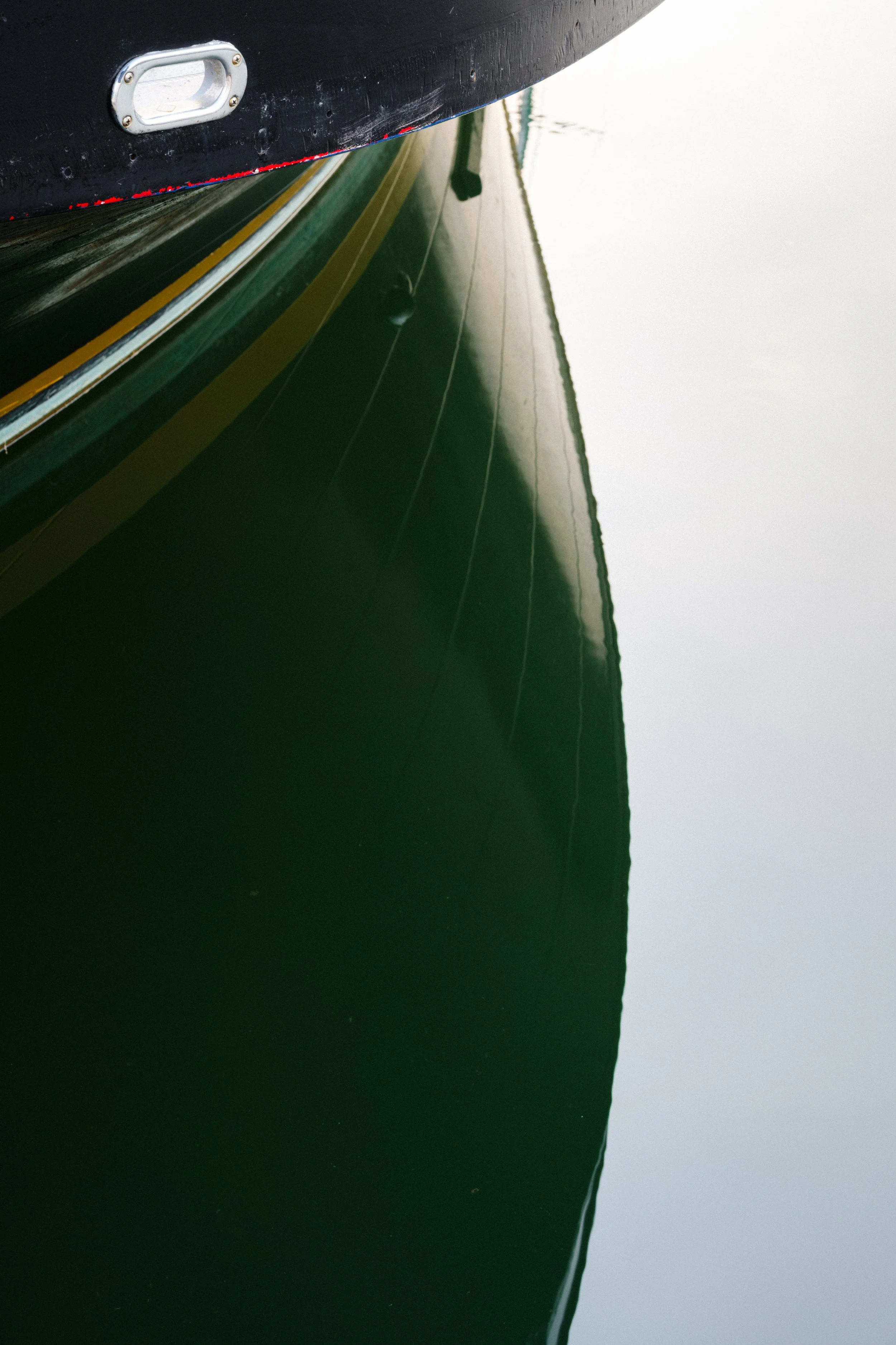 Close-up view of the side of a boat reflected in the water, showing the hull, a metallic cleat, and a thin yellow stripe on the boat's body.