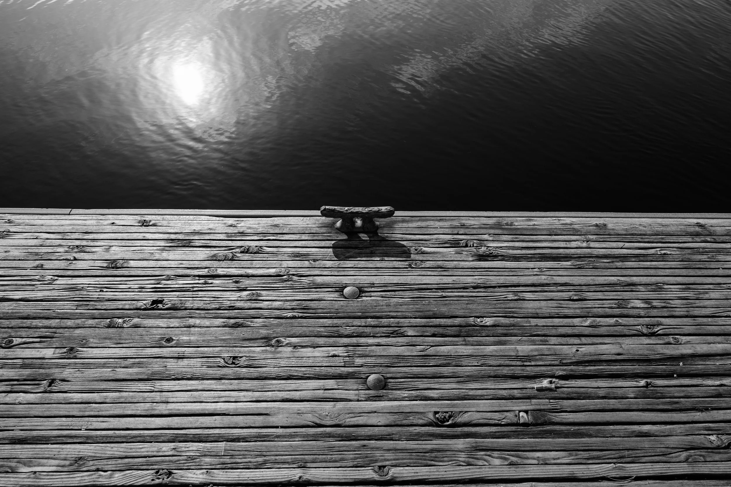 Black and white photo of a wooden dock with metal cleat and two floating balls, overlooking a calm body of water with sunlight reflection.