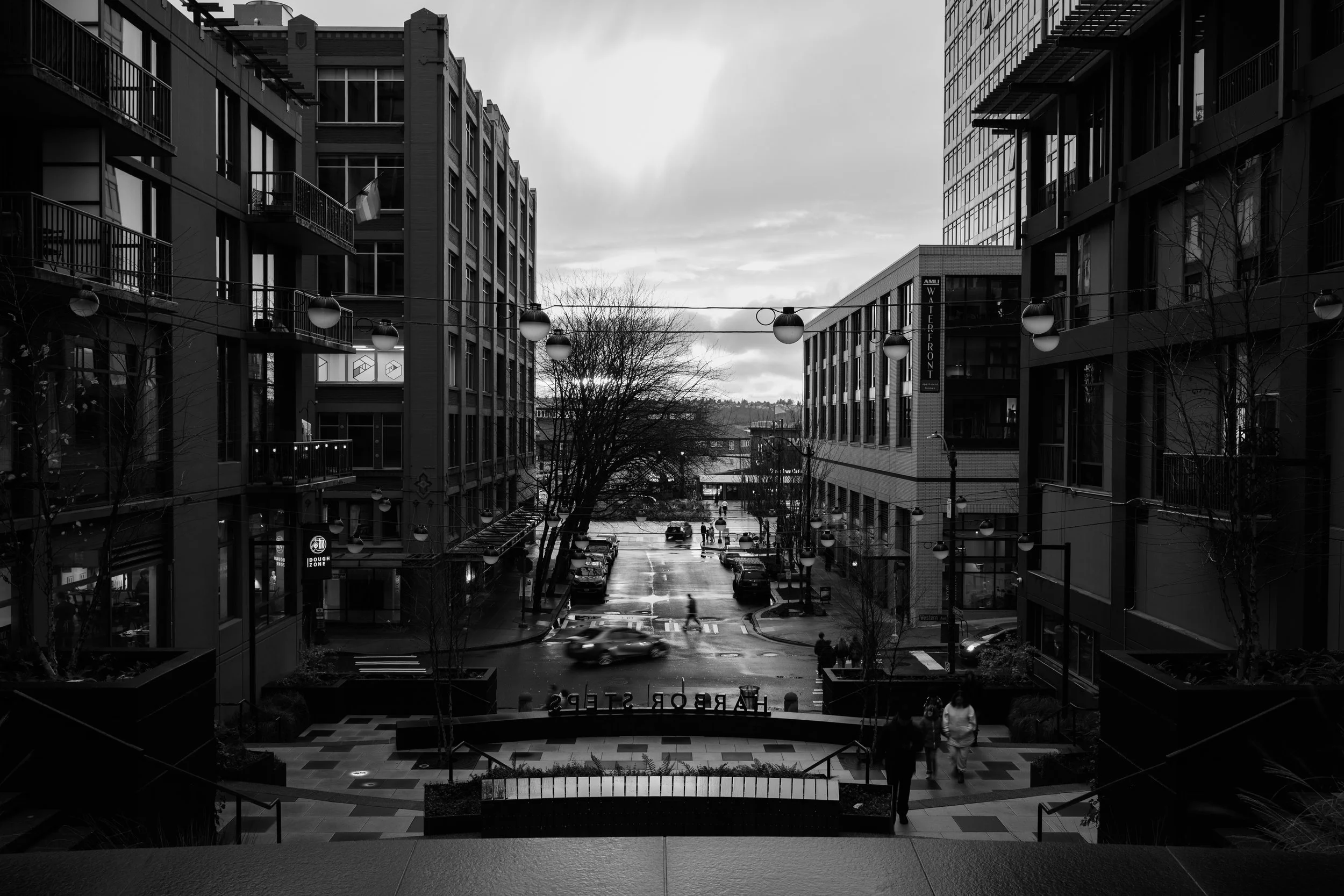 Black and white photo of an urban street scene with modern buildings on either side. The street has cars parked along the curb and a few pedestrians walking. Overhead, there are decorative globe streetlights strung across the street. The sky is cloud