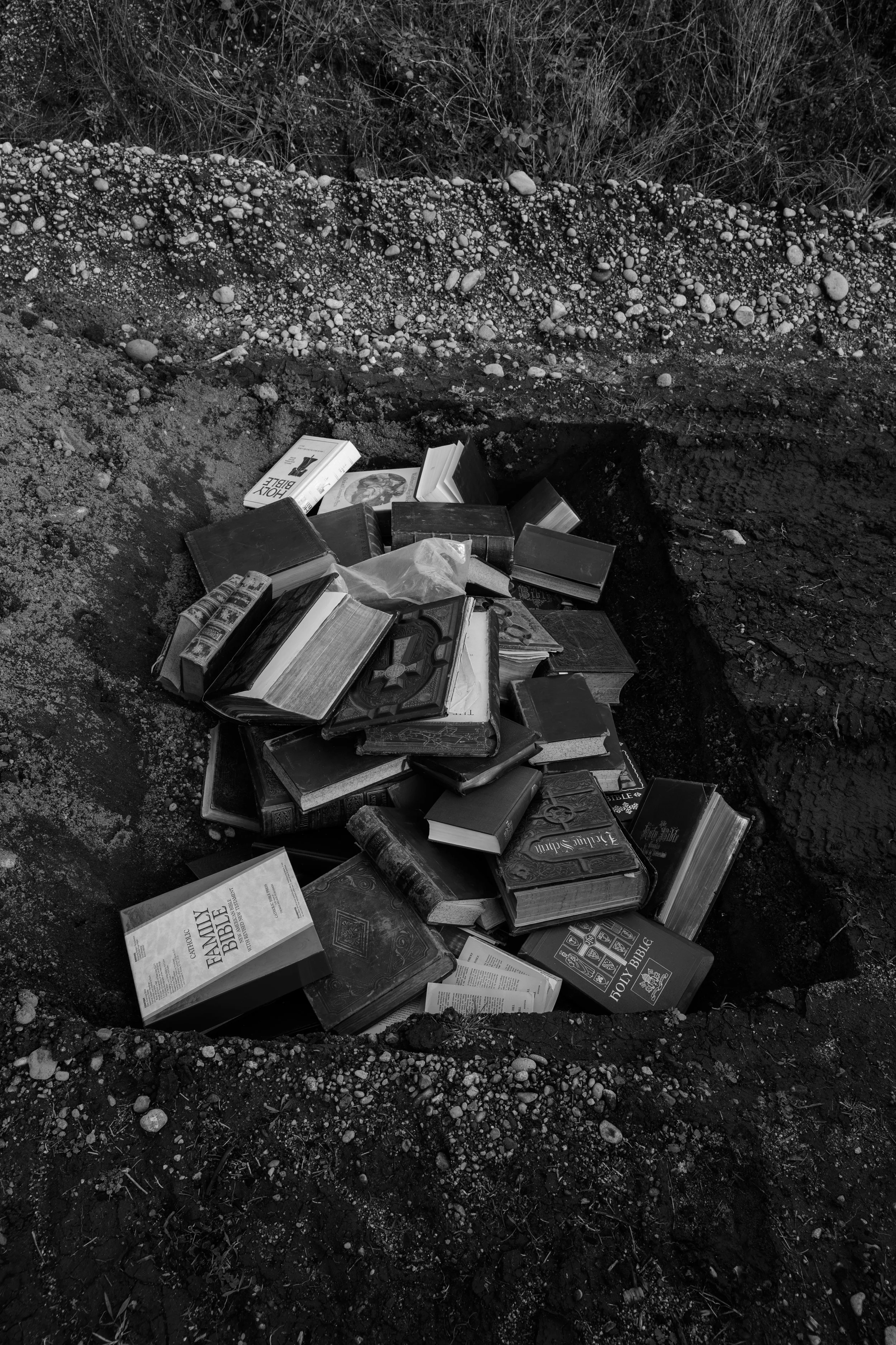 Pile of open and closed books, including a Bible, in a hole in the ground.