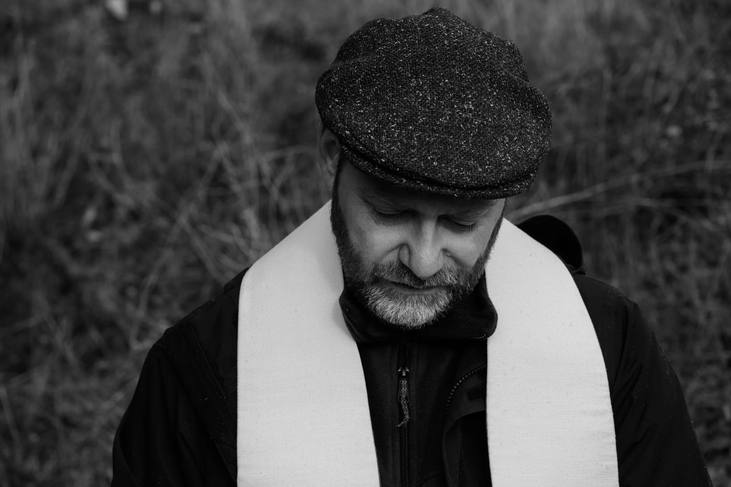 A man with a beard and mustache, wearing a textured flat cap, looking down, outdoors with blurred natural background, in black and white.