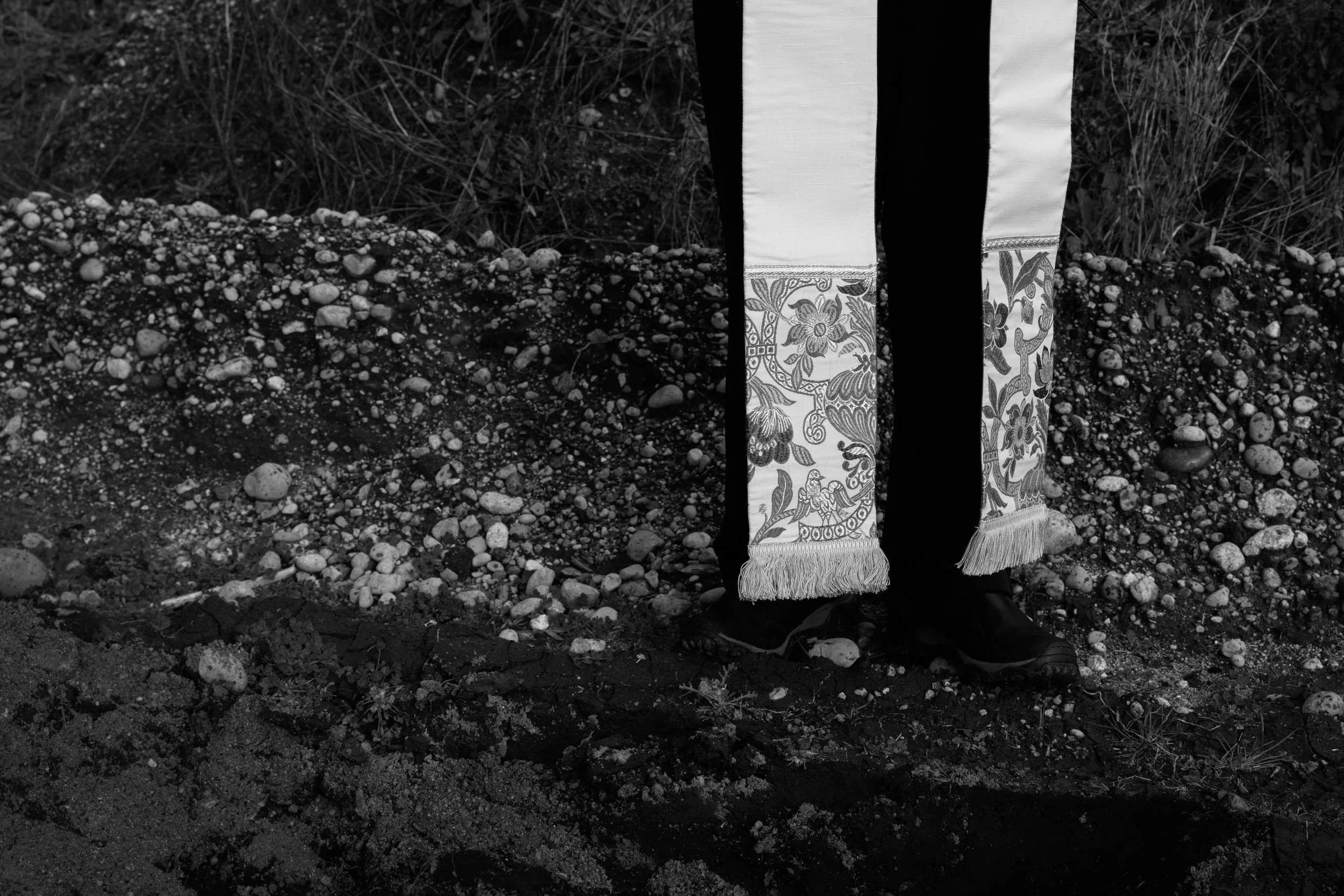 Lower half of a person standing on rocky ground, wearing dark pants and black shoes; the person has a decorative white garment with floral embroidery and fringe at the bottom.