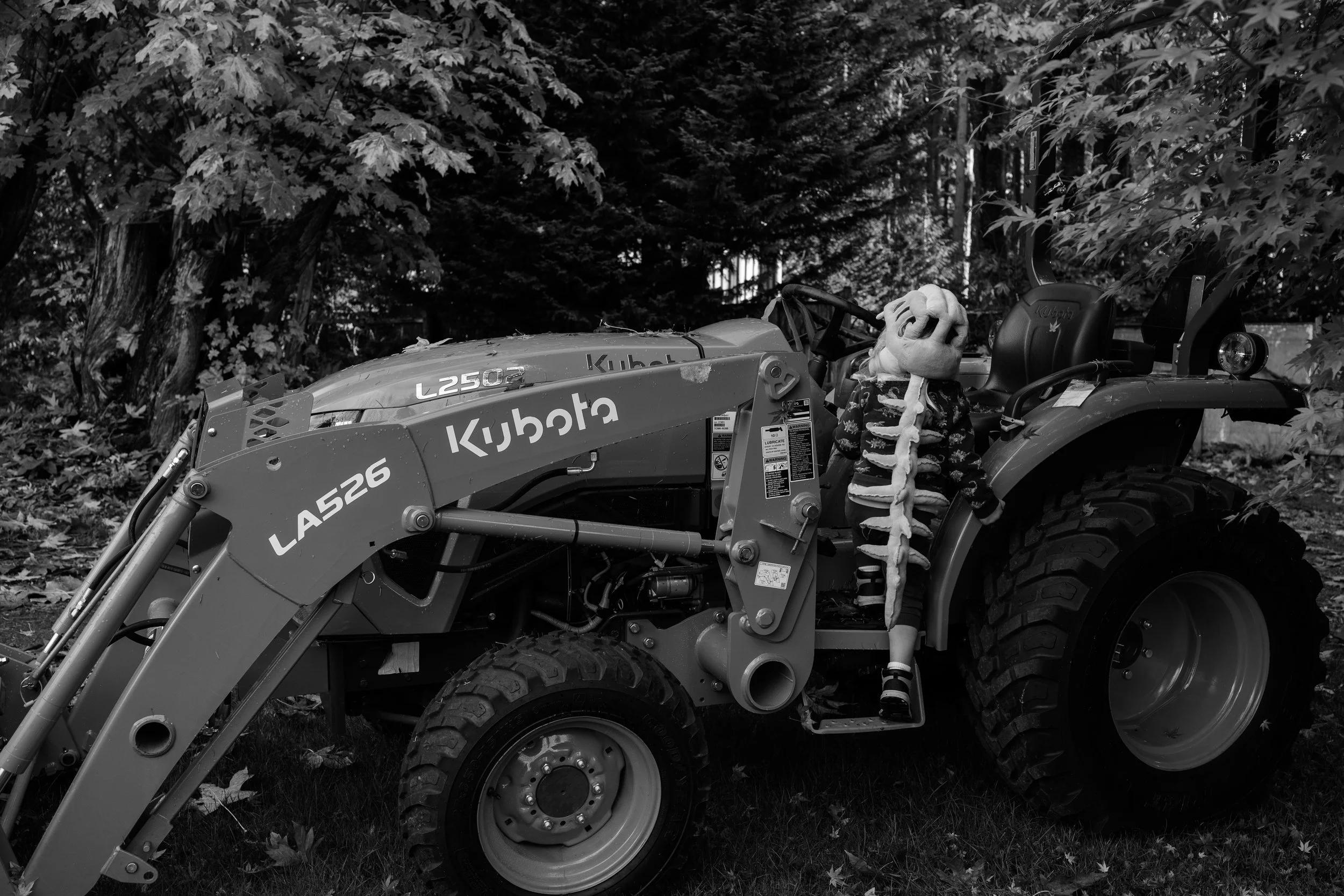 A black and white photo of a child in a skeleton mask standing next to a tractor with a front loader attachment, surrounded by trees and leaves.
