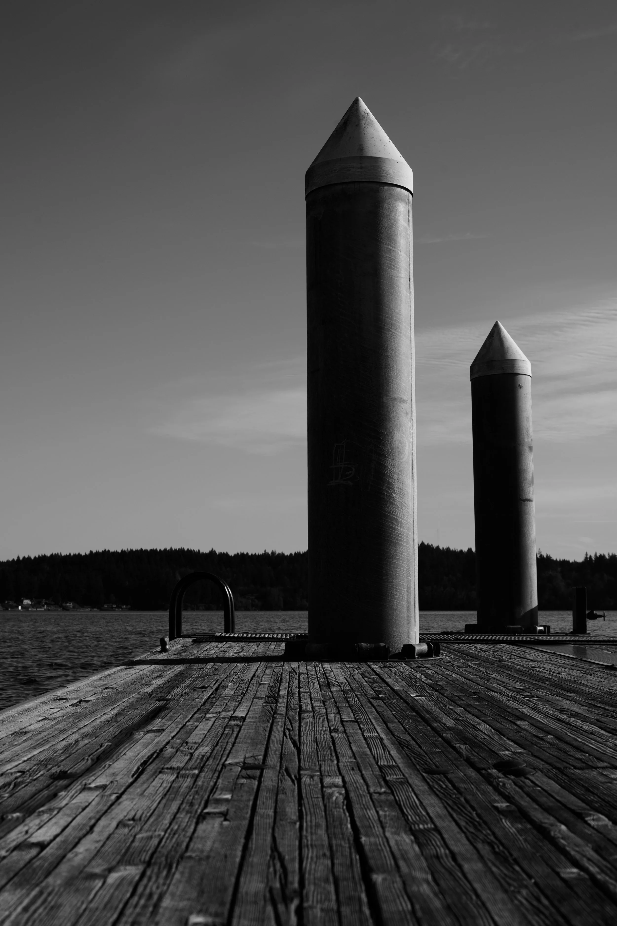 Black and white photo of a wooden dock with two large cylindrical metal posts with pointed tops, beside a body of water, with a distant tree-covered shoreline in the background.