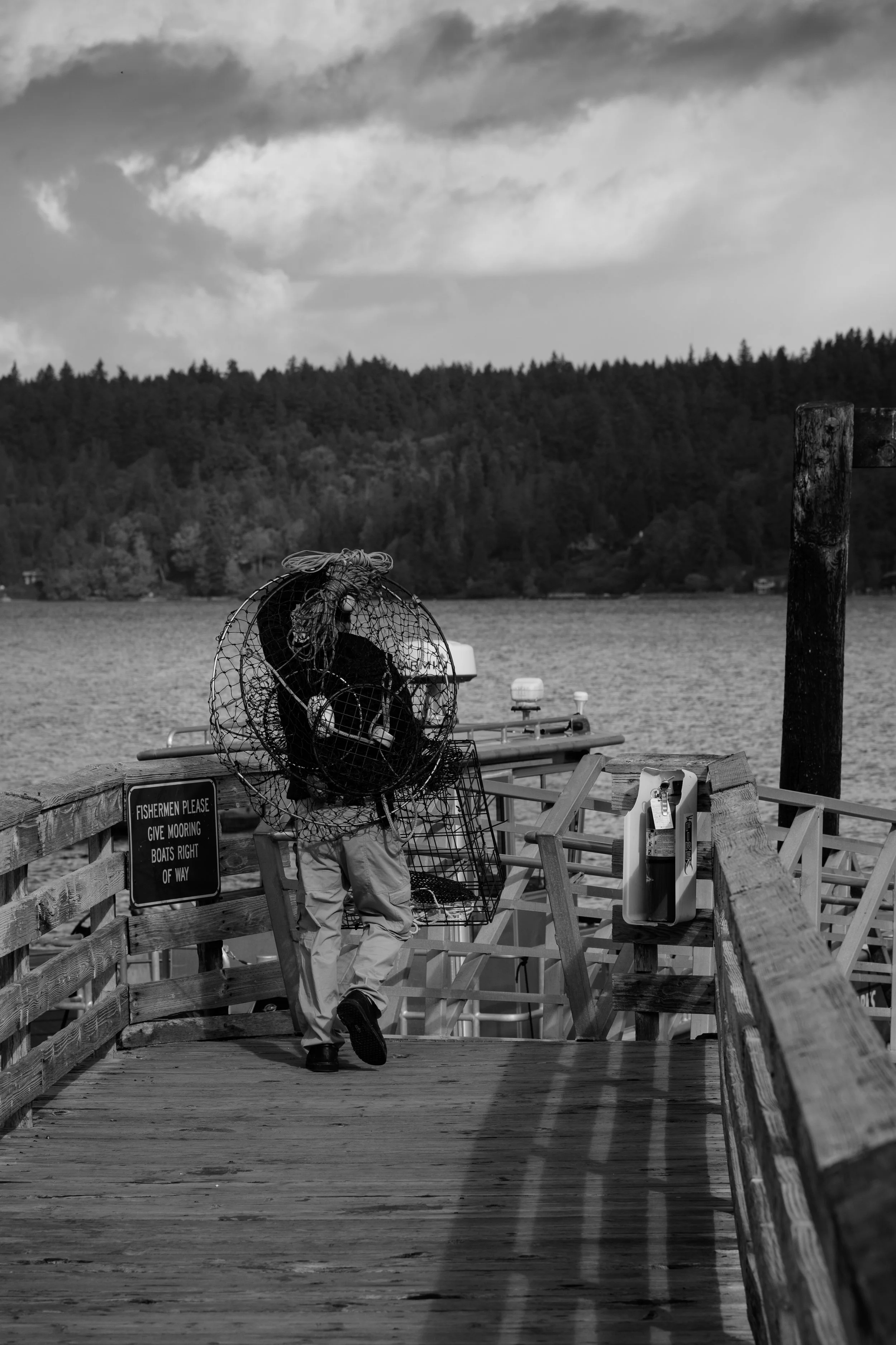 A person walking away on a wooden dock carrying a large crab trap or cage, with water and a forested shoreline in the background, on a cloudy day.